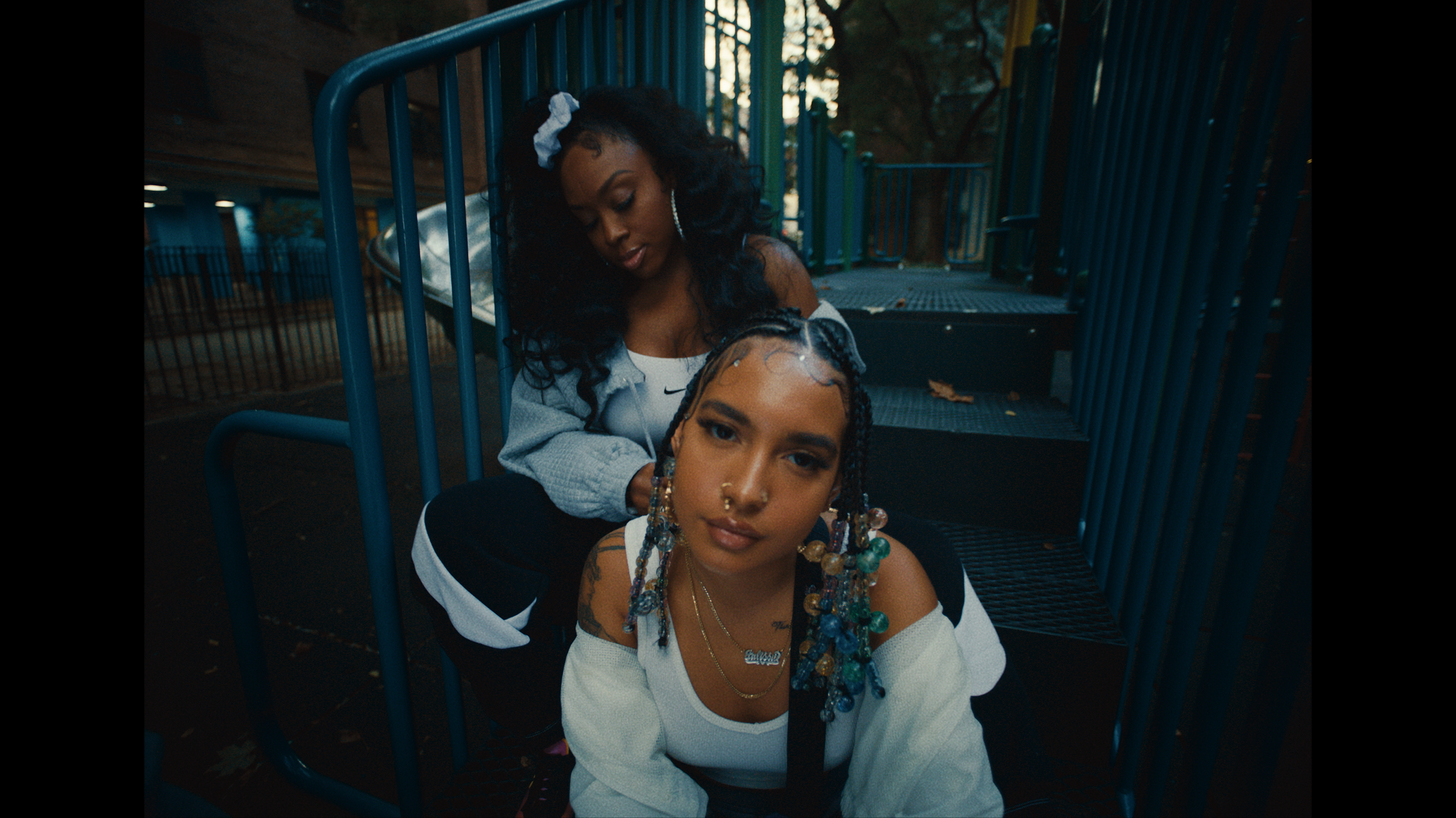 Two young women sitting on steps at a playground. One with braids and colorful beads, the other with curly black hair and a white scrunchie, both dressed casually in athletic wear.