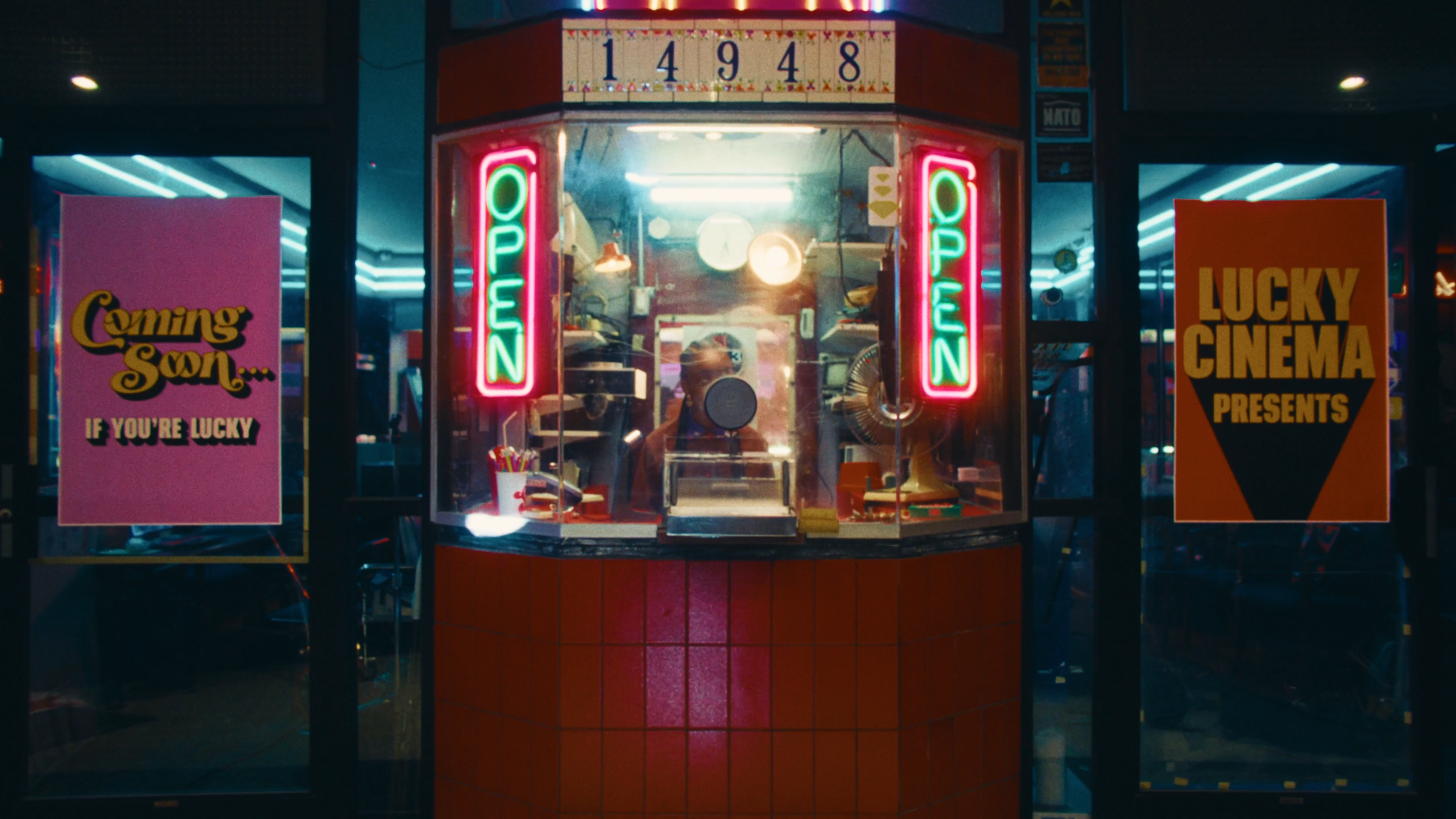 Neon signs in a small shop window, with one sign reading 'OPEN' and another 'COMING SOON... IF YOU'RE LUCKY'. The shop has red tiled exterior, and inside on the wall, there is a date '14948' displayed.