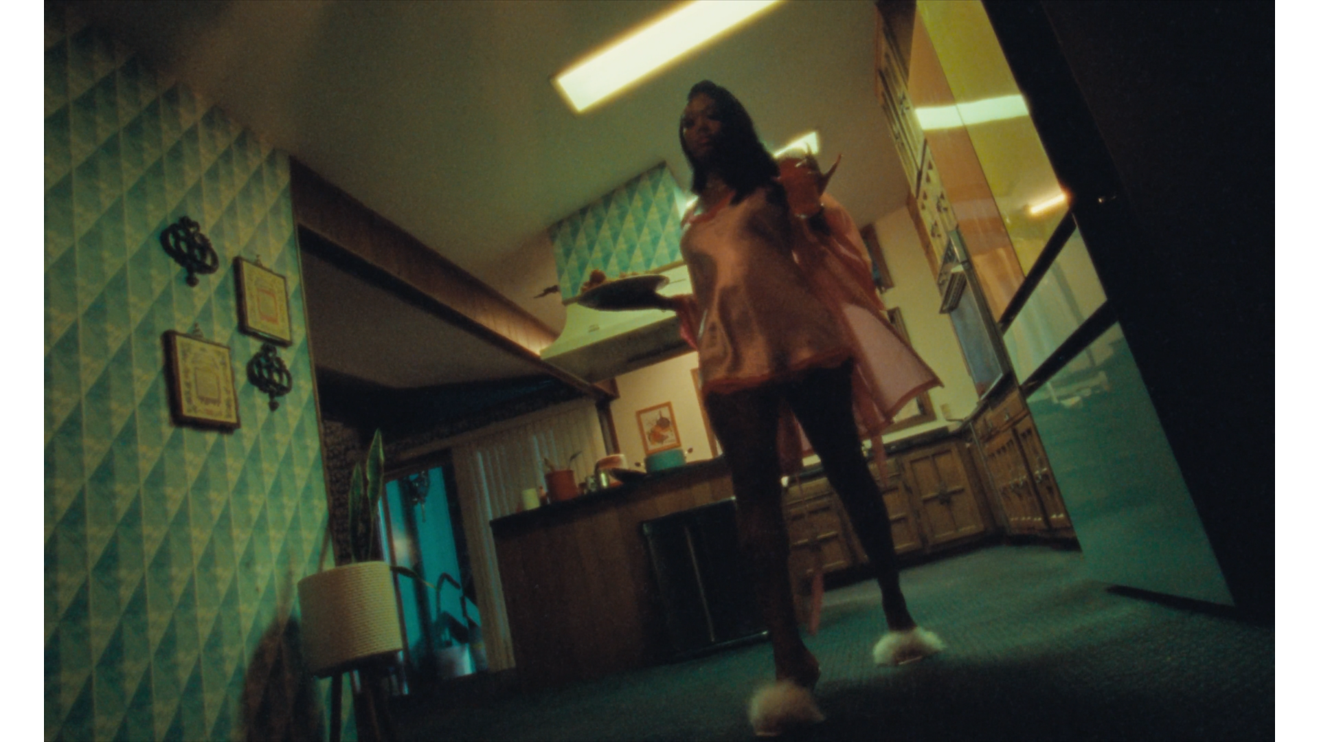 A woman walking in a kitchen holding a plate, with retro decor and wooden cabinets.