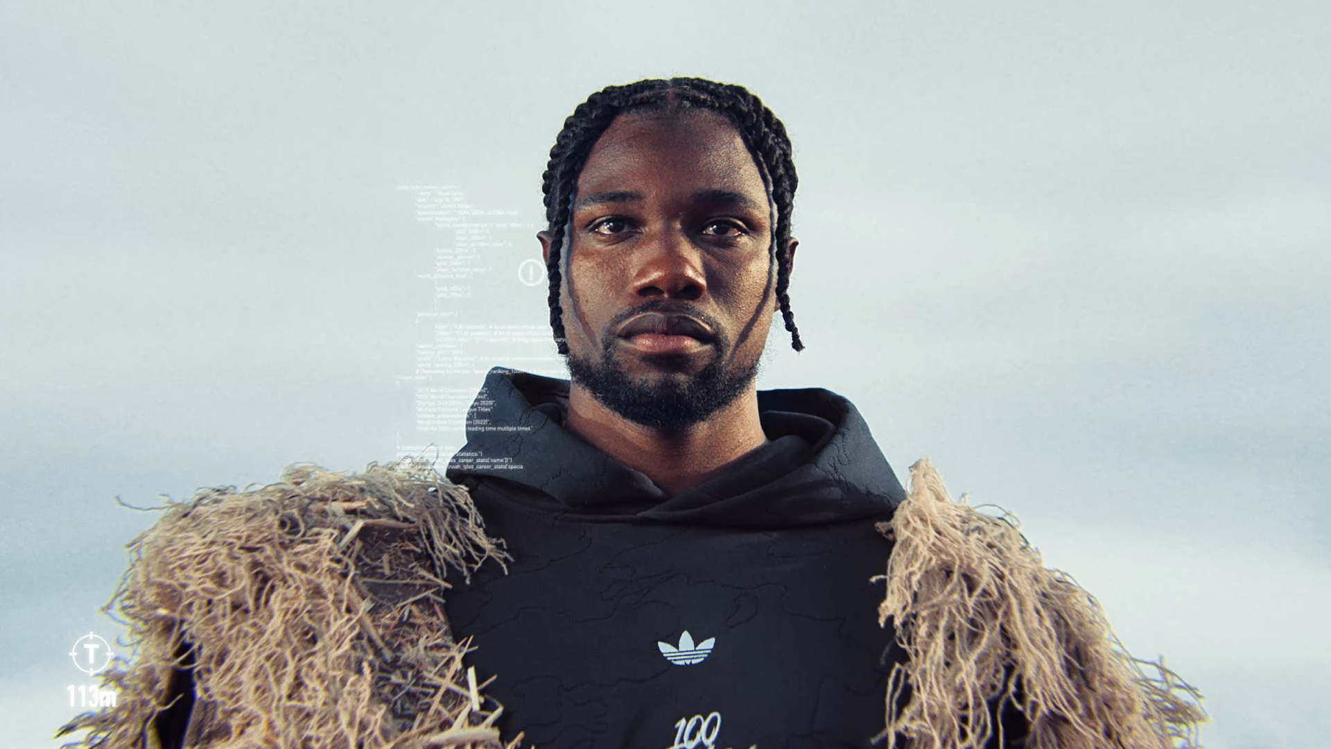 A man with dreadlocks and a beard wearing a black hoodie with a small logo, and a beige, shaggy, textured jacket, standing against a gray sky.