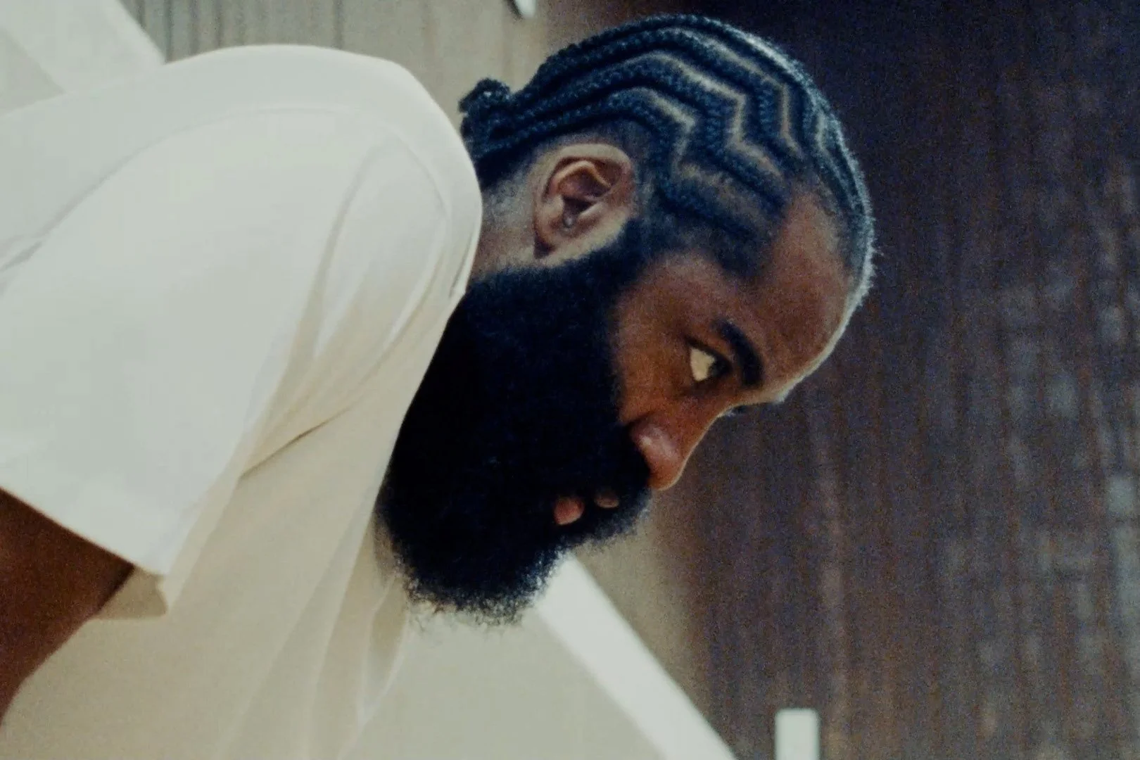 A person with blue braided hair and a dark beard wearing a white shirt, looking down.