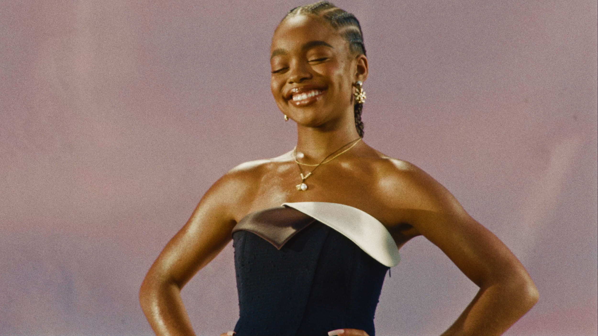 A woman with dark skin and braided hair wearing a strapless black dress with white and brown accents, smiling with eyes closed, against a plain background.