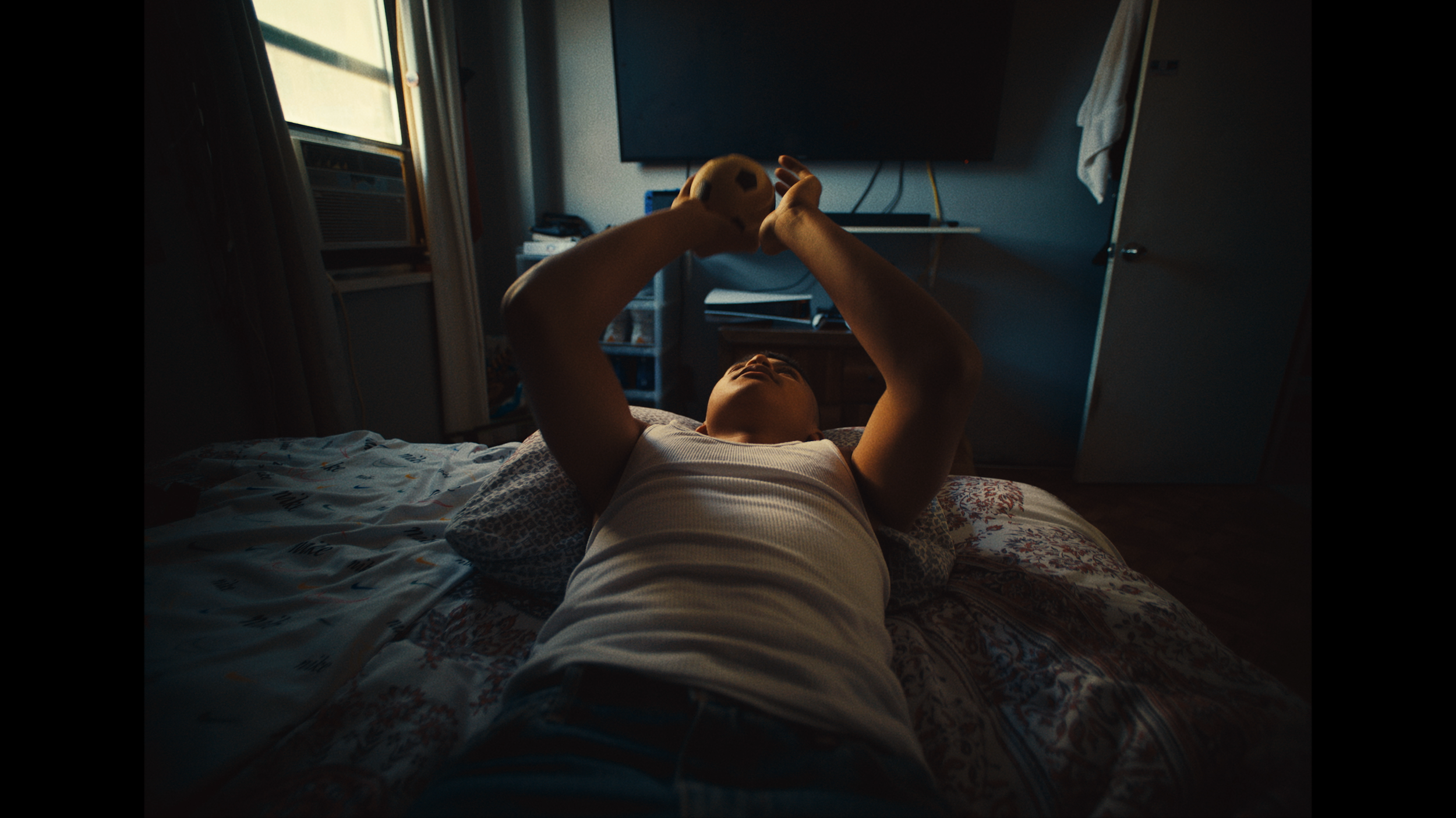 A person lying on a bed playing with a soccer ball in a dimly lit bedroom.