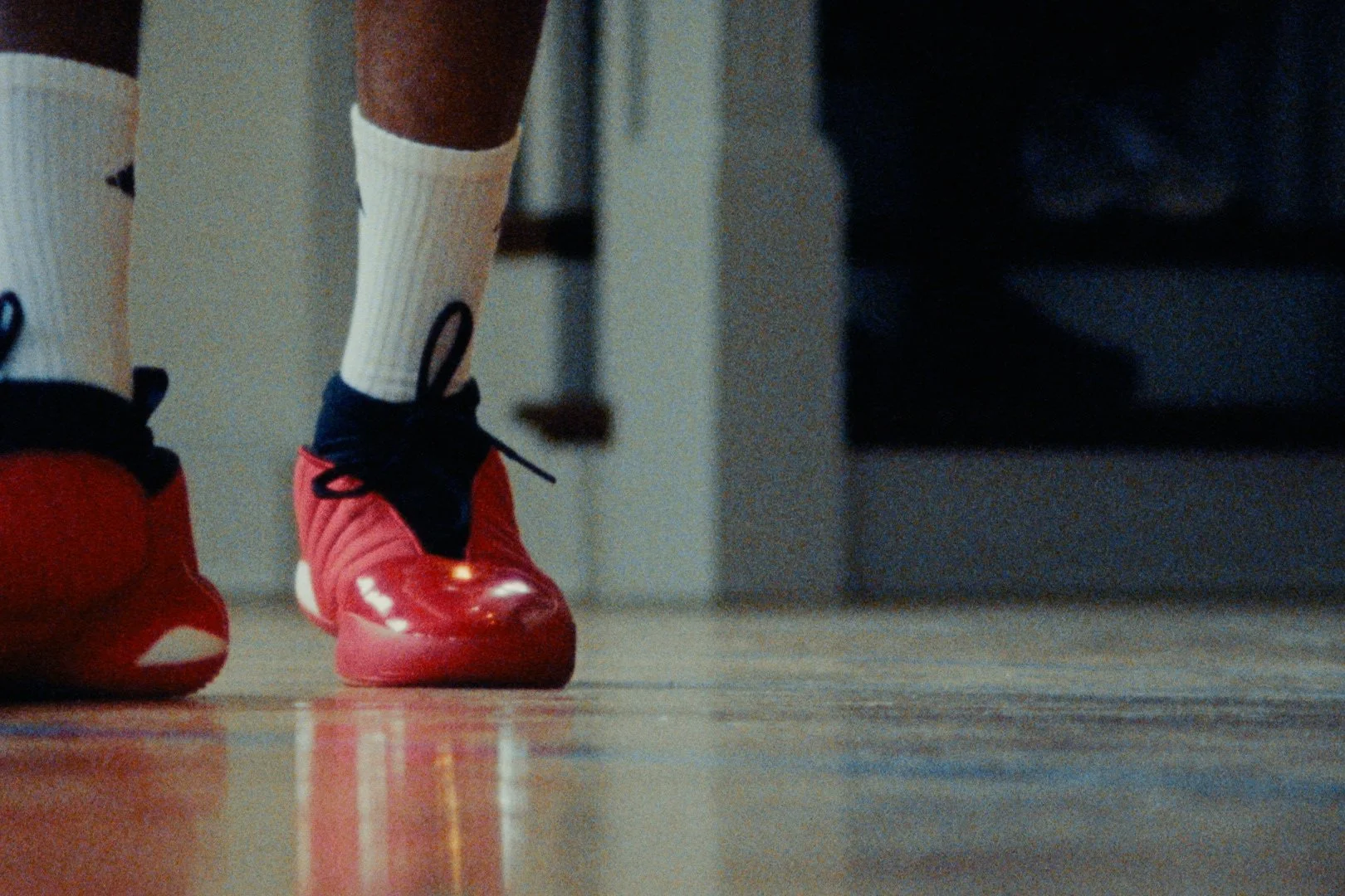 Close-up of a person's legs wearing white sports socks and red athletic shoes, walking on a wooden floor.
