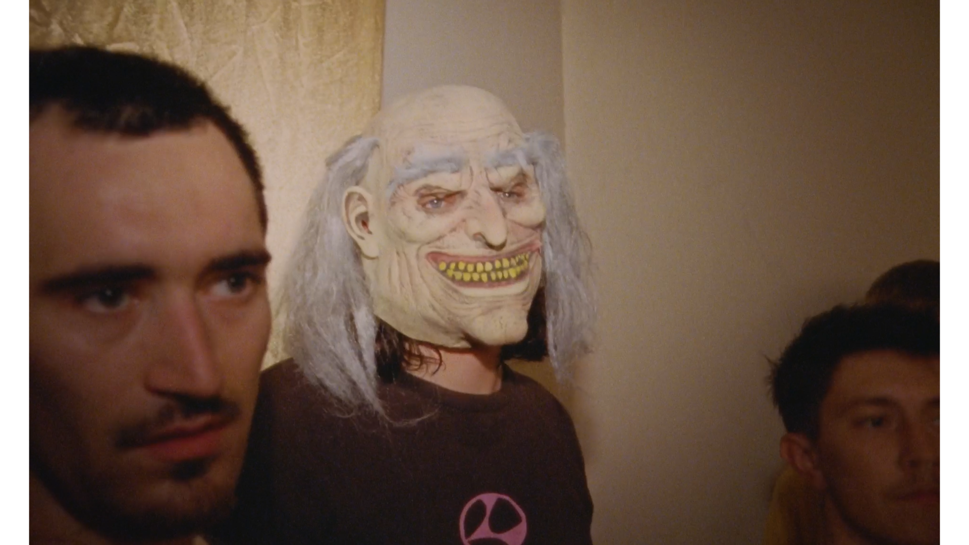Three people sitting indoors, with the person in the center wearing a creepy mask resembling an old, wrinkled face with long white hair, exhibiting a wide toothy grin.