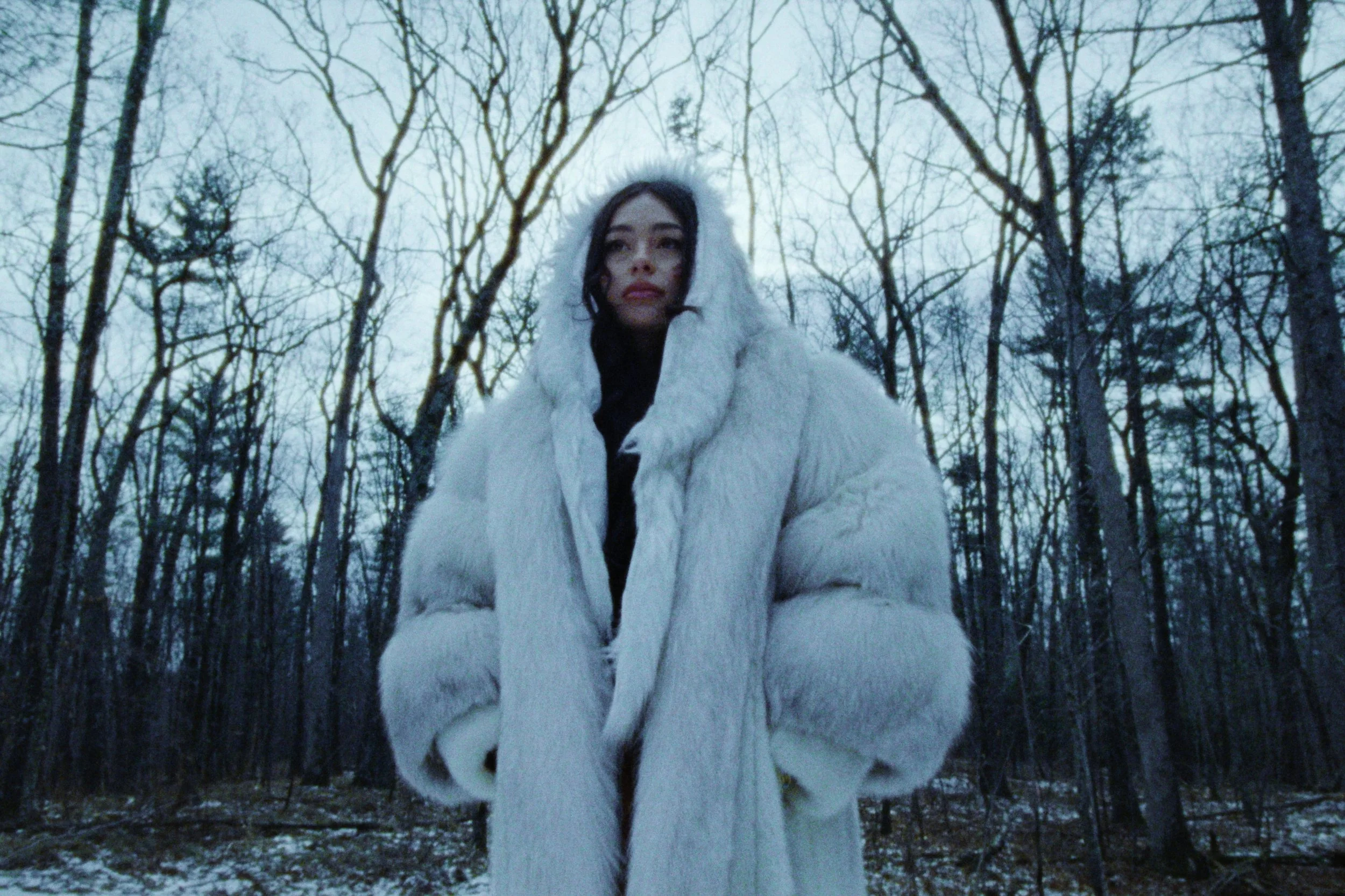 A woman in a white fur coat standing outdoors in a leafless forest during winter.