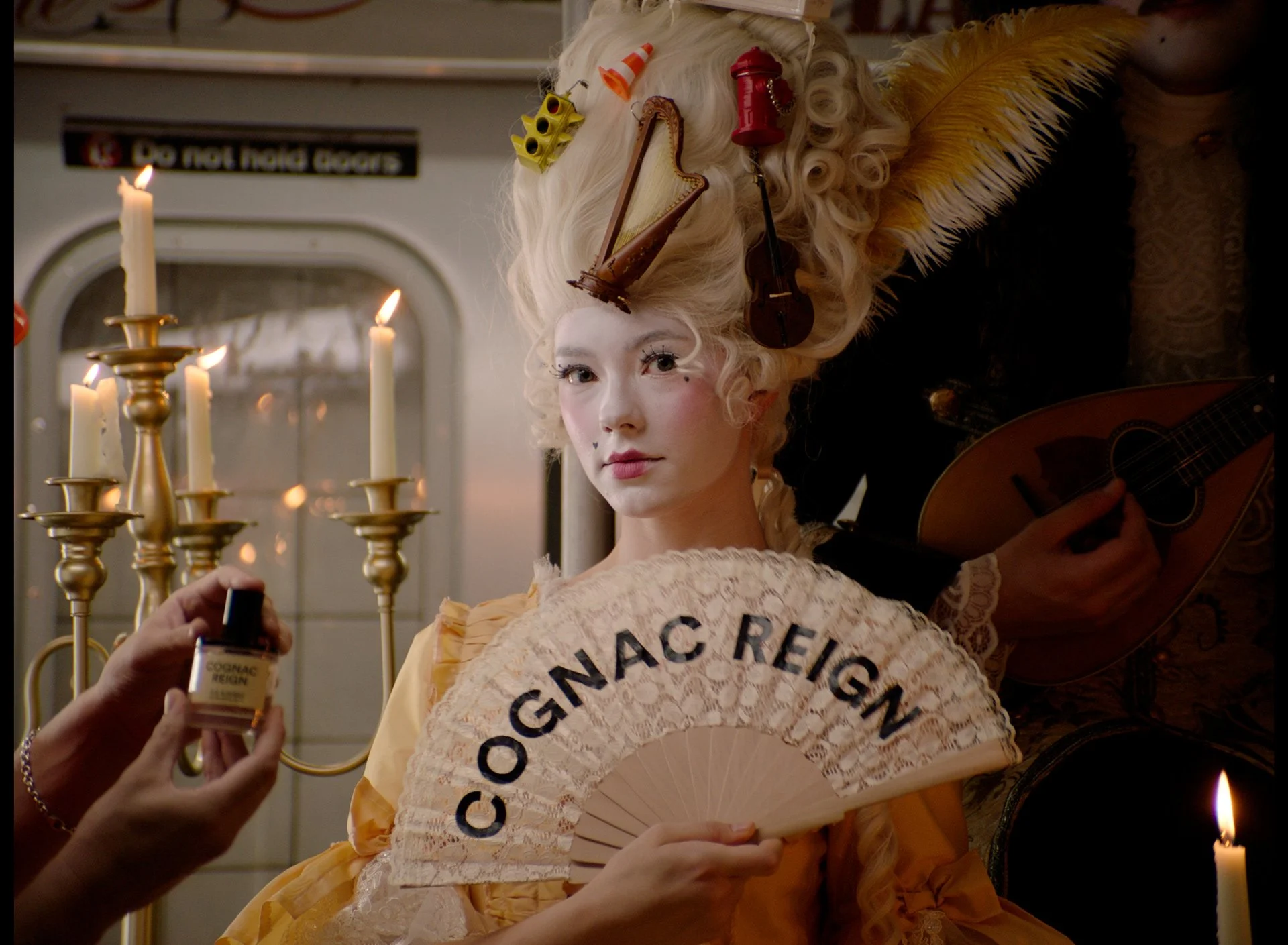 A woman in elaborate 18th-century costume, holding a fan labeled "Cognac Reign," sits in front of lit candles and a mirror, with a man beside her playing a guitar.