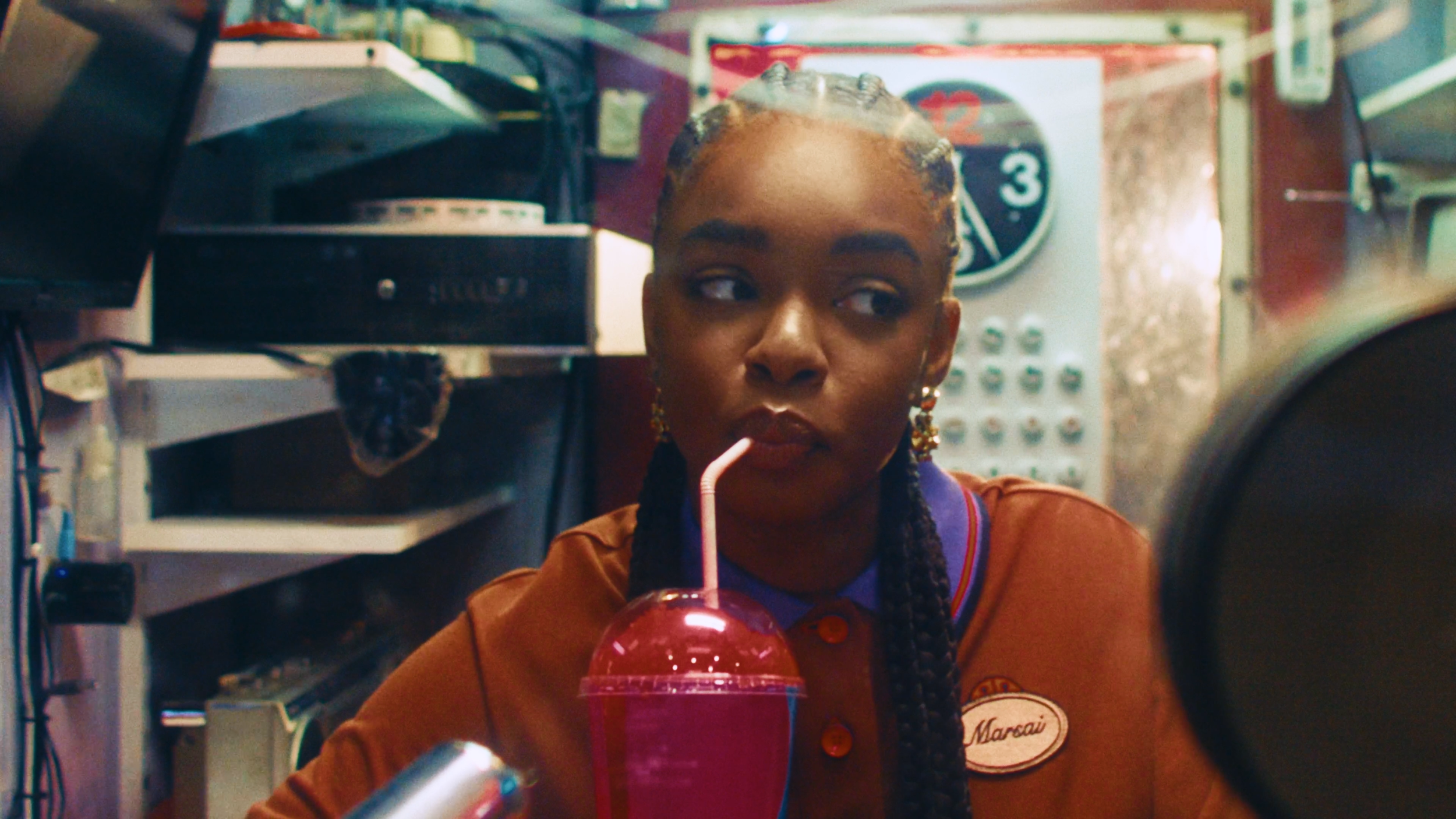 A young woman with braided hair and gold earrings inside a convenience store, sipping a pink milkshake through a straw. She wears a brown uniform with a nametag that reads 'Marsai.' Behind her, shelves and a payphone are visible.