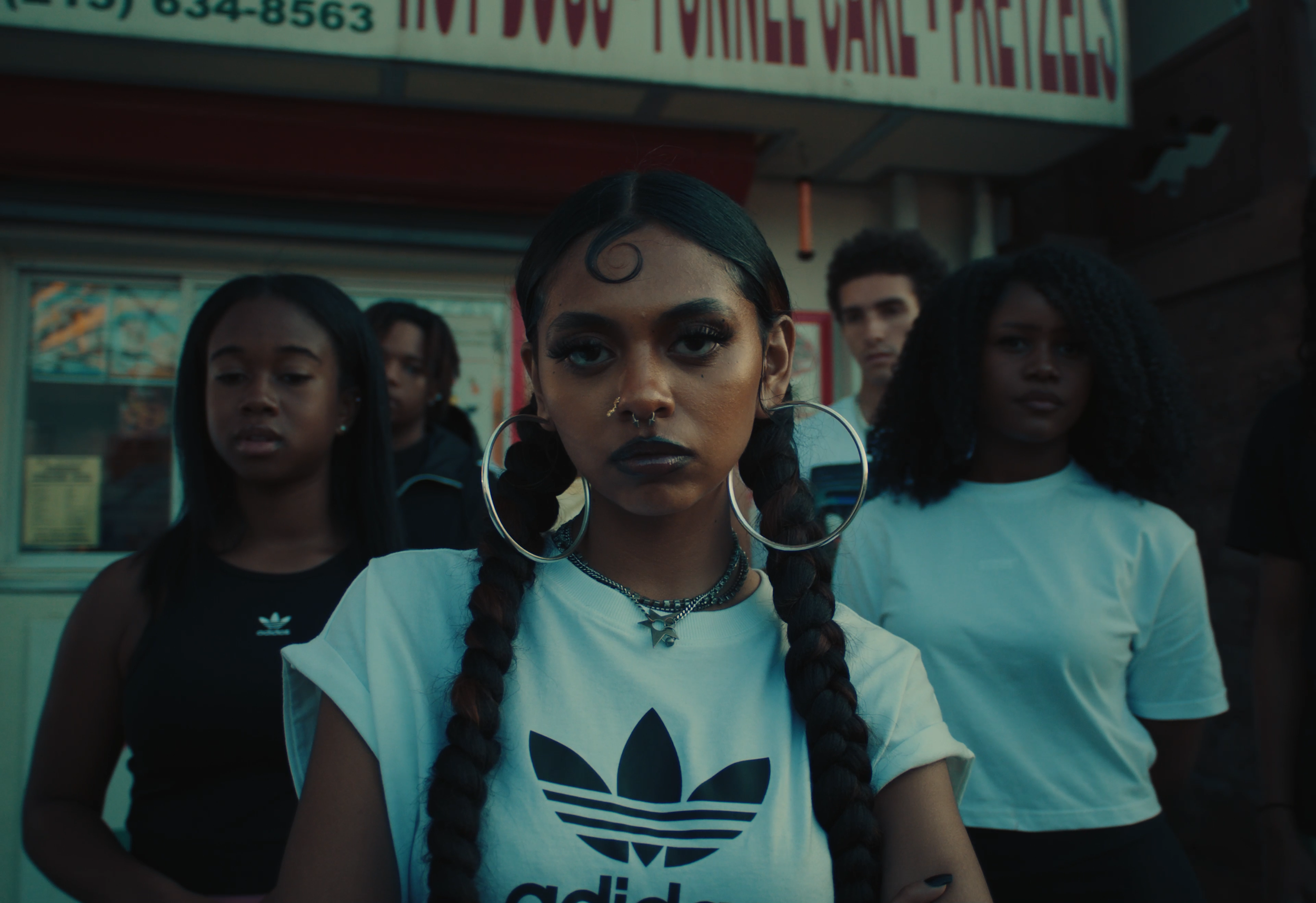 A group of diverse young people standing outdoors, with the focus on a girl with braided hair, large hoop earrings, and dark lipstick, wearing a white Adidas shirt, in front of a fast food restaurant.