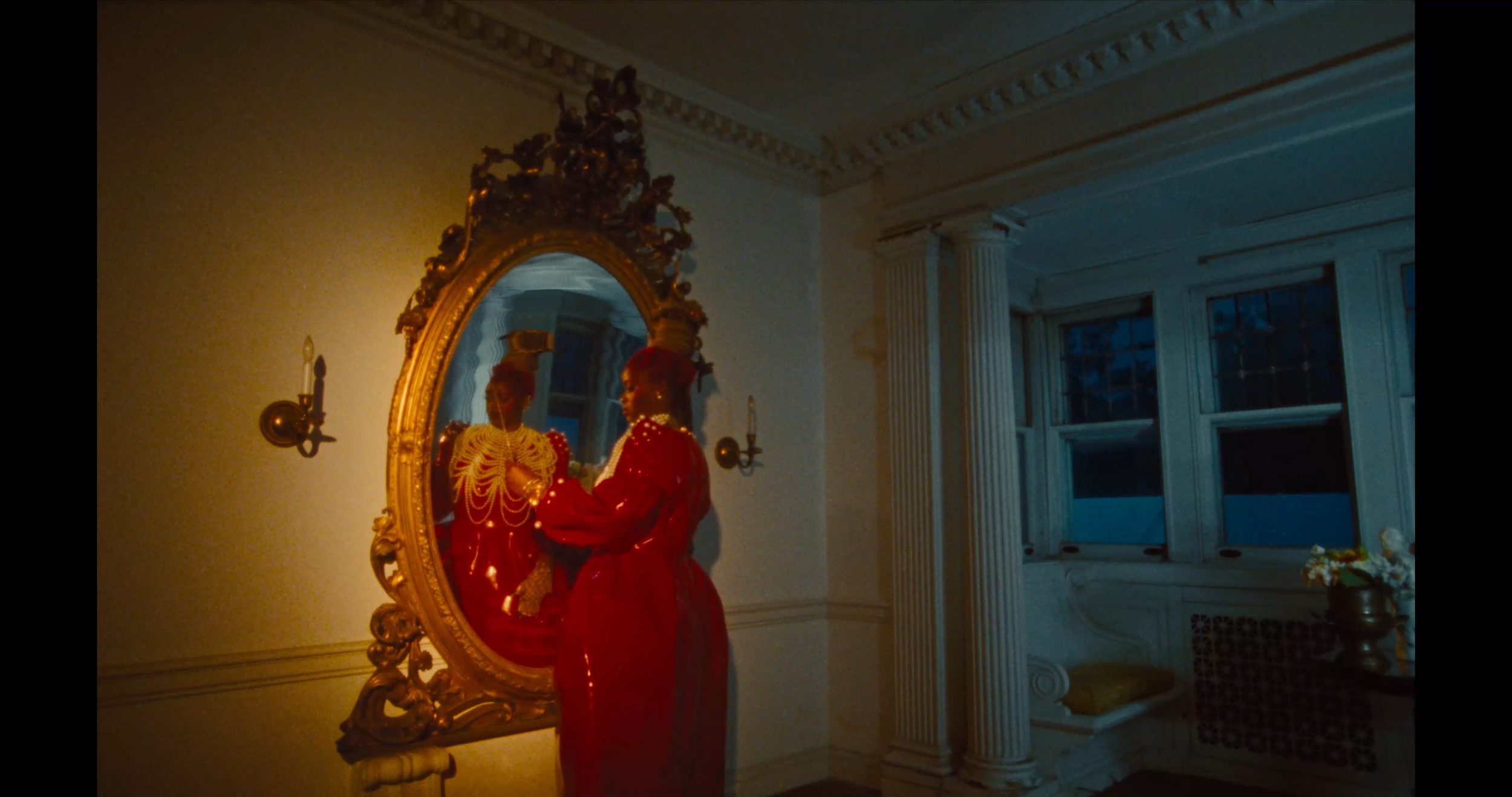 A woman in a red dress standing in front of an ornate gold-framed mirror, looking at her reflection. The room has classical architectural features including white columns, crown molding, and a bench by the window. There are candles on the wall next t