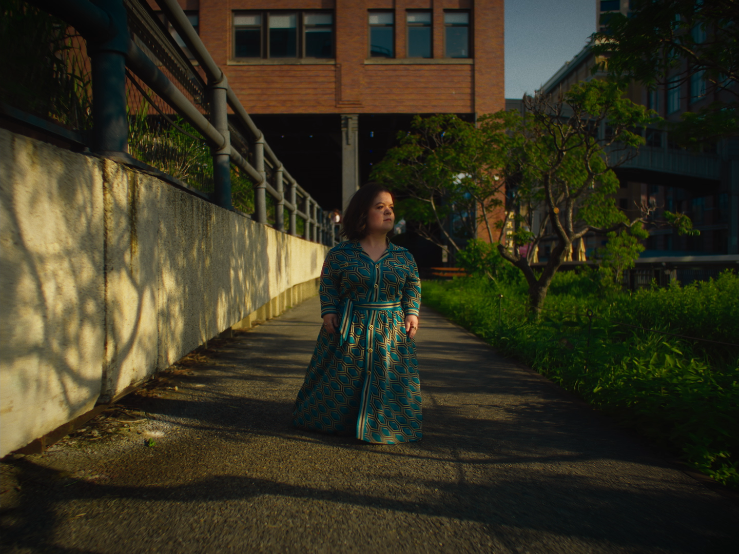 A woman in a patterned dress walks along a city sidewalk during late afternoon or early evening, with buildings, greenery, and shadows in the background.