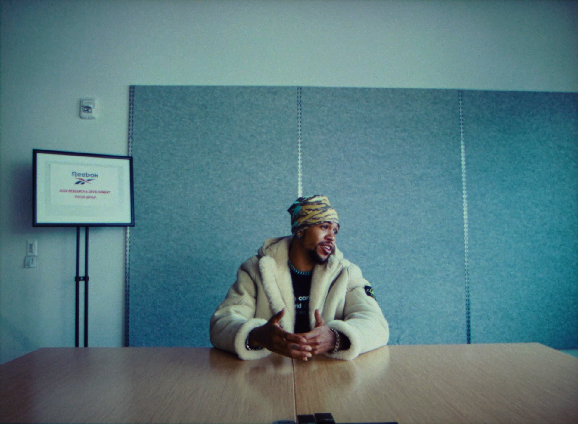 A man sitting at a wooden table in a modern conference room, wearing a beige fuzzy jacket and a colorful headscarf, gesturing with his hands while speaking. There is a framed Reebok sign and a gray acoustic panel wall in the background.