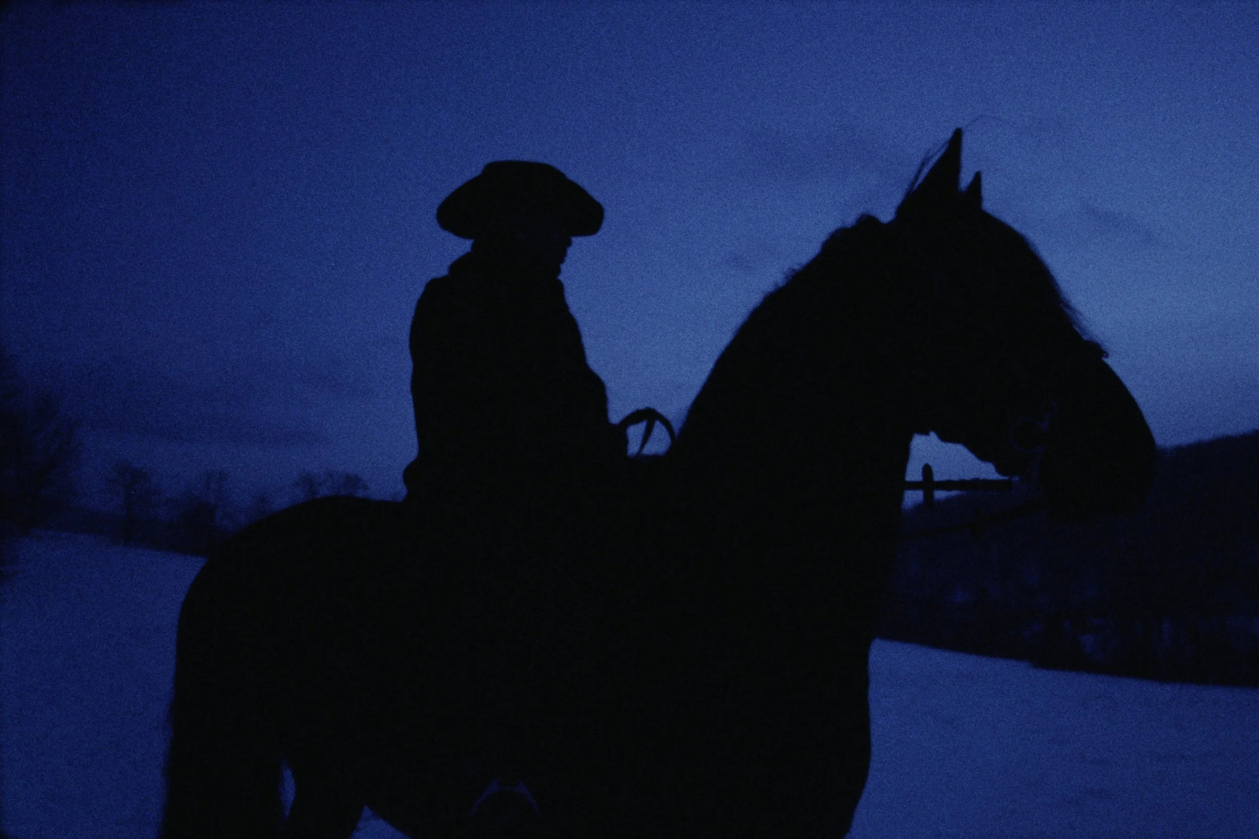 Silhouette of a cowboy riding a horse against a dark blue evening sky.