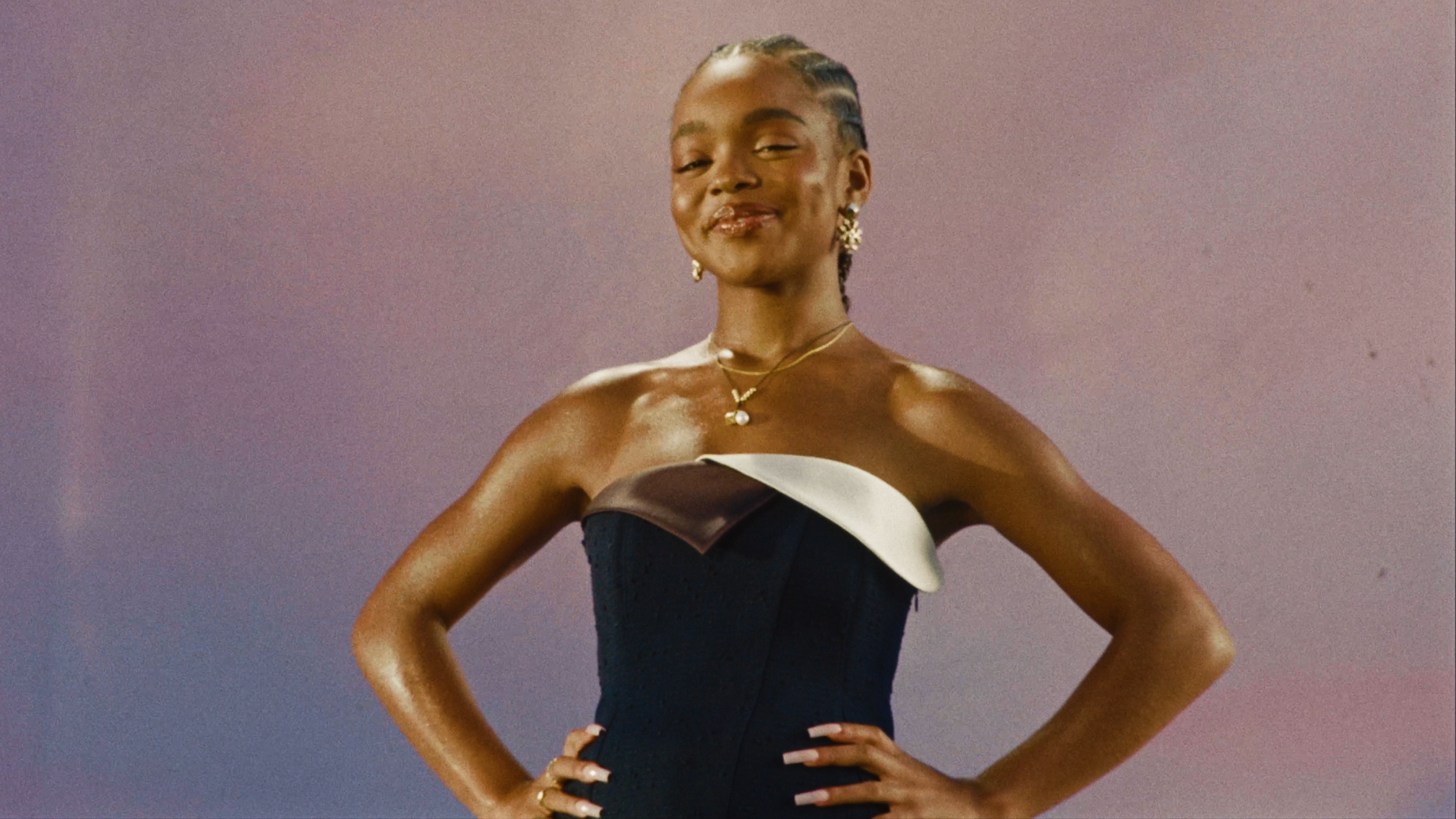 A woman with dark skin, wearing a strapless black and white dress, standing with her hands on her hips against a gradient pink background. She has jewelry, including earrings and necklaces, and is smiling confidently.