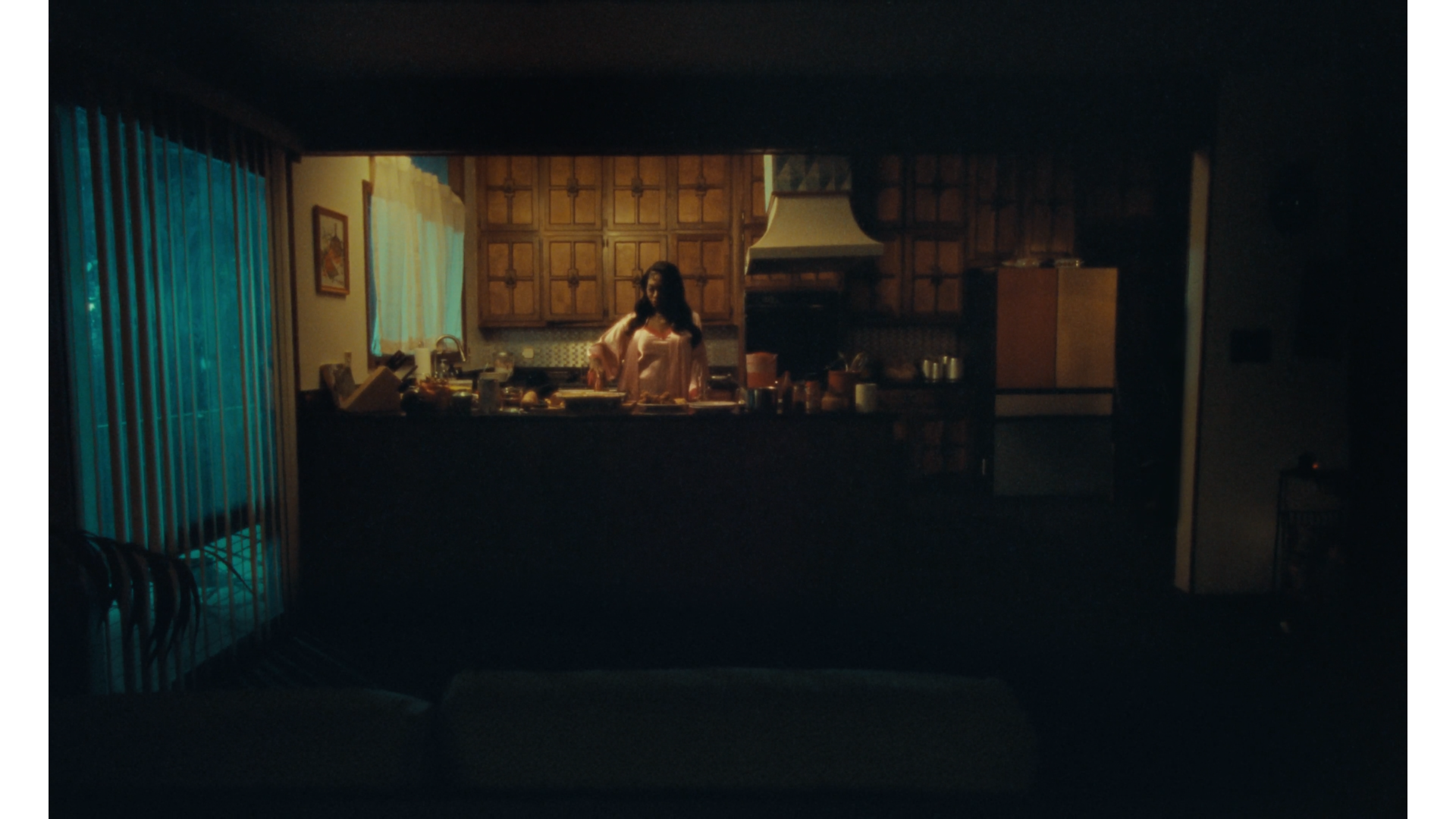 A woman in pajamas standing in a dimly lit kitchen, seen through a window from a living room with a dark silhouette in the foreground.