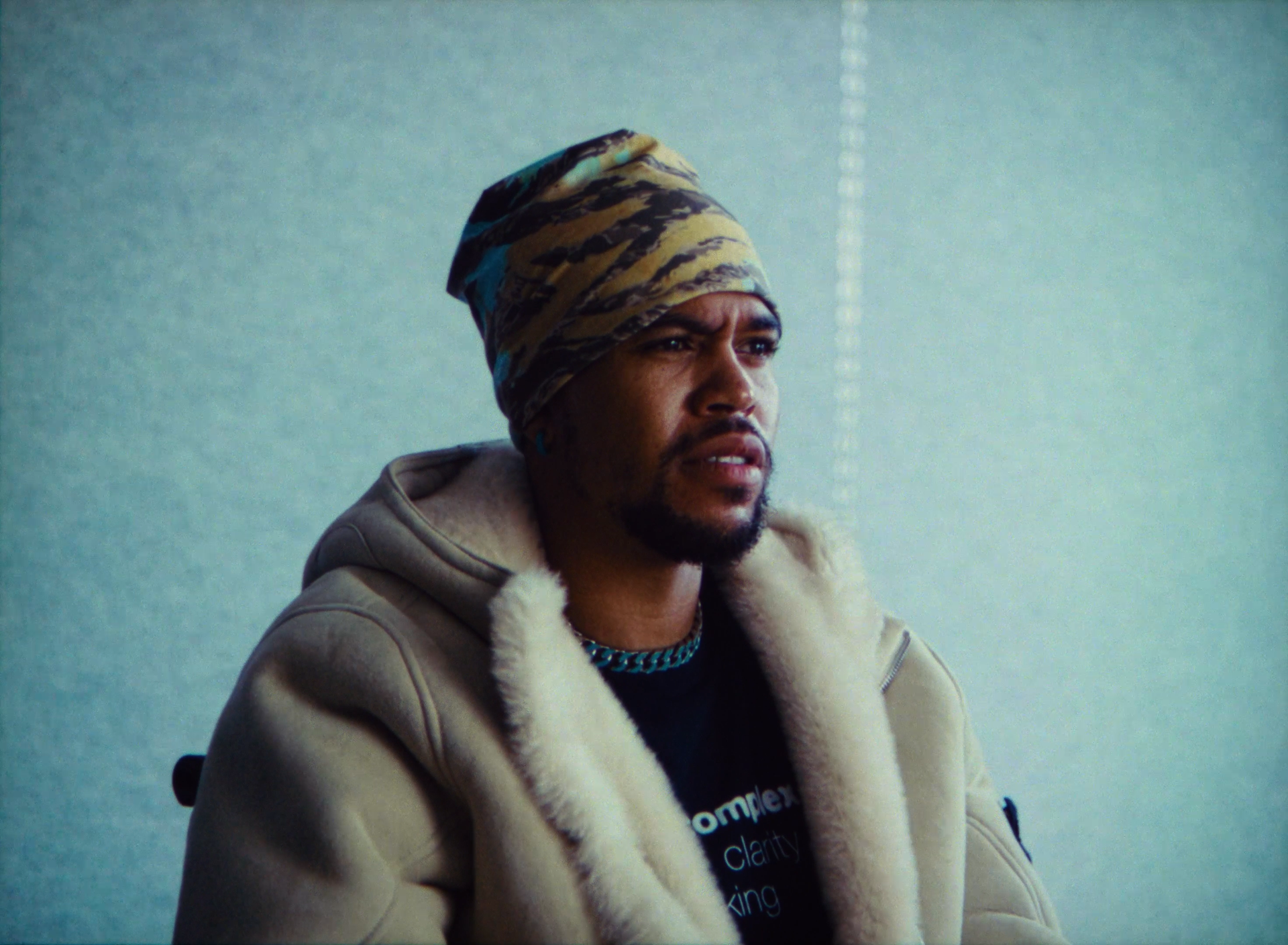 A man with a serious expression, wearing a tan jacket with a fur collar, a patterned headscarf, a chain necklace, and a black T-shirt, sitting indoors against a plain background.