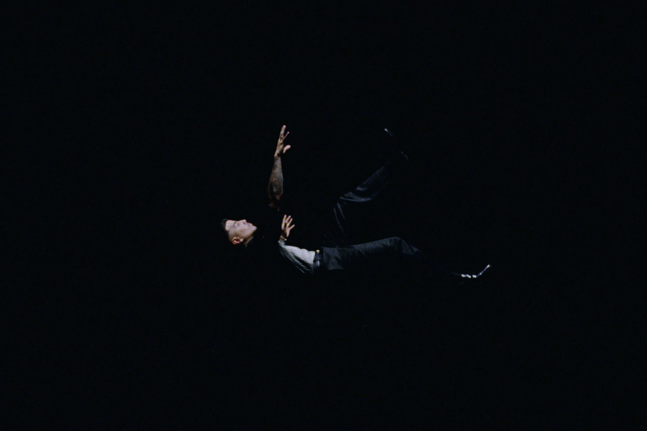 A man floating in a dark space, wearing dark clothing, with his arms and legs extended and his head tilted back.