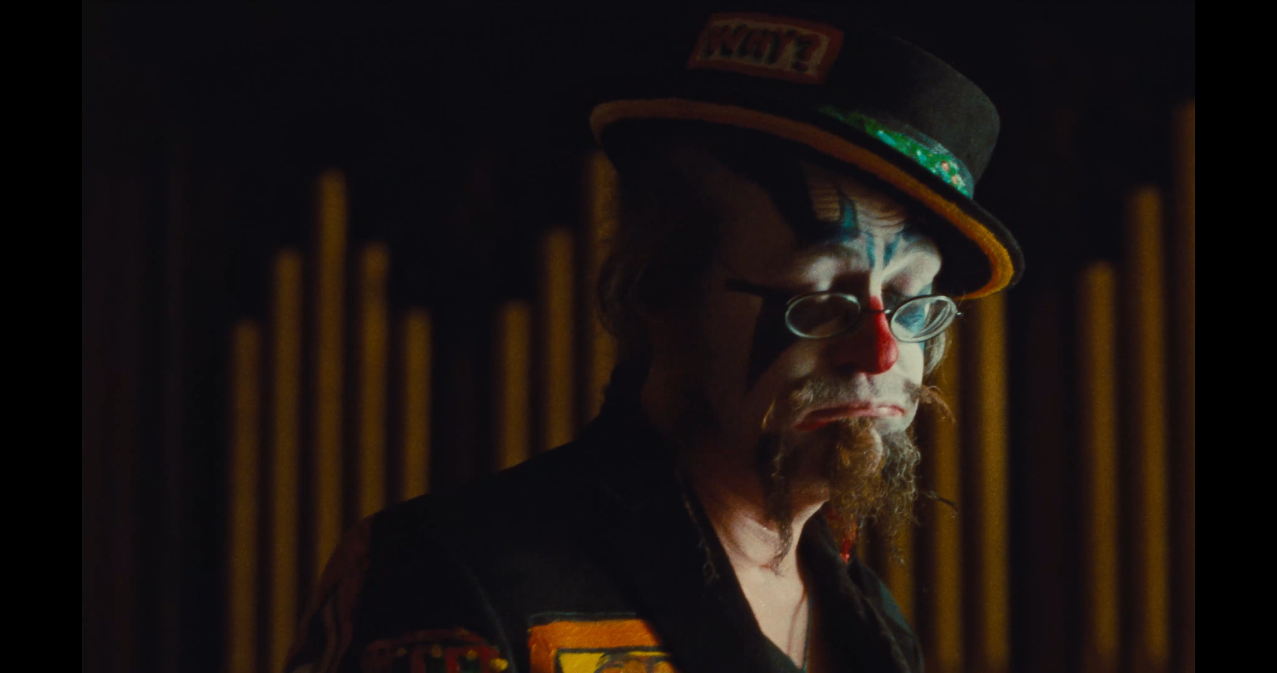 A person dressed as a clown with a painted face, wearing a hat, glasses, and a colorful jacket, with a dark background.