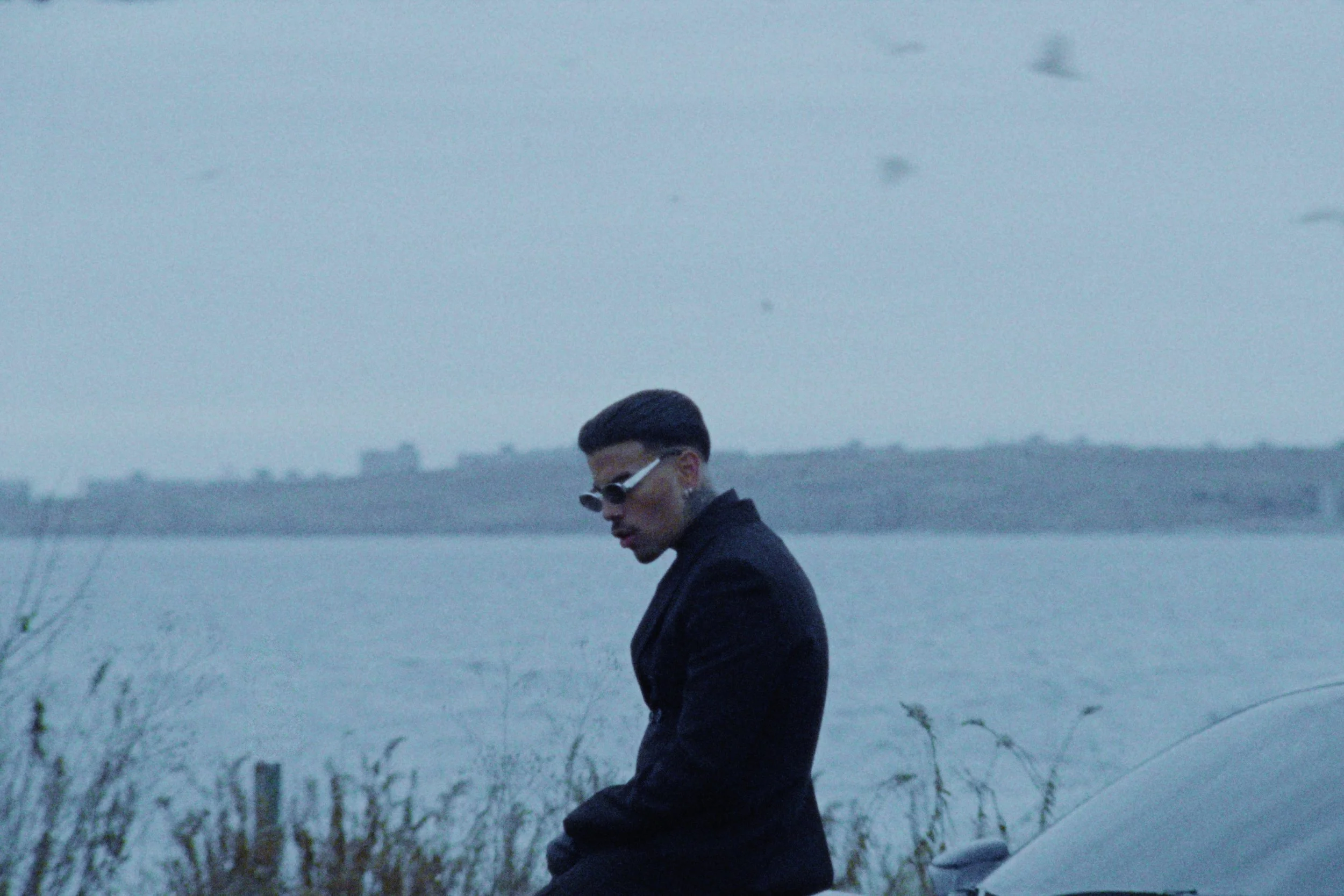 A man with dark hair, wearing sunglasses and a dark coat, walking near a body of water with a foggy or overcast sky.