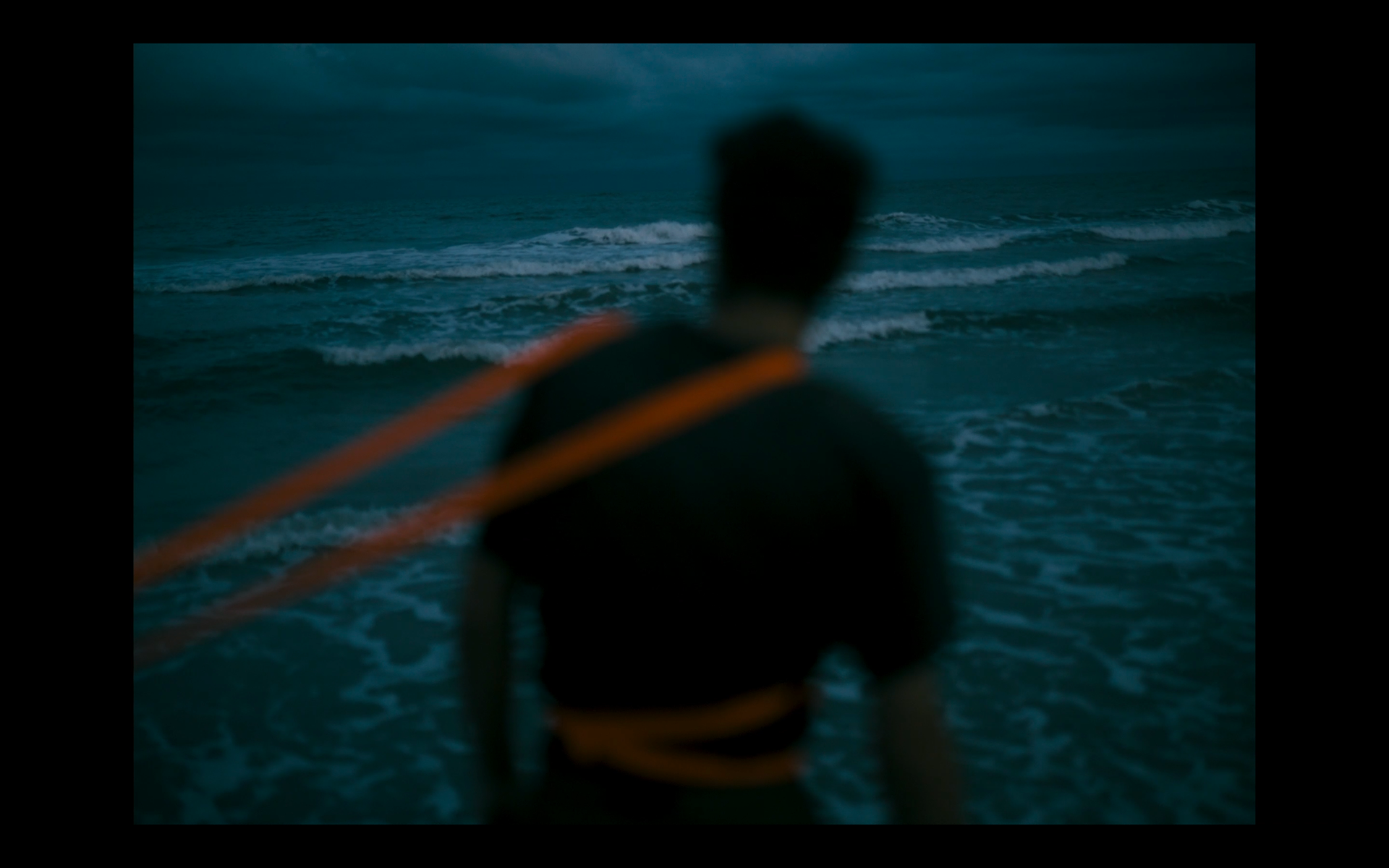 Blurred person standing on a beach at dusk, facing the ocean with waves, with orange ropes around their shoulder.