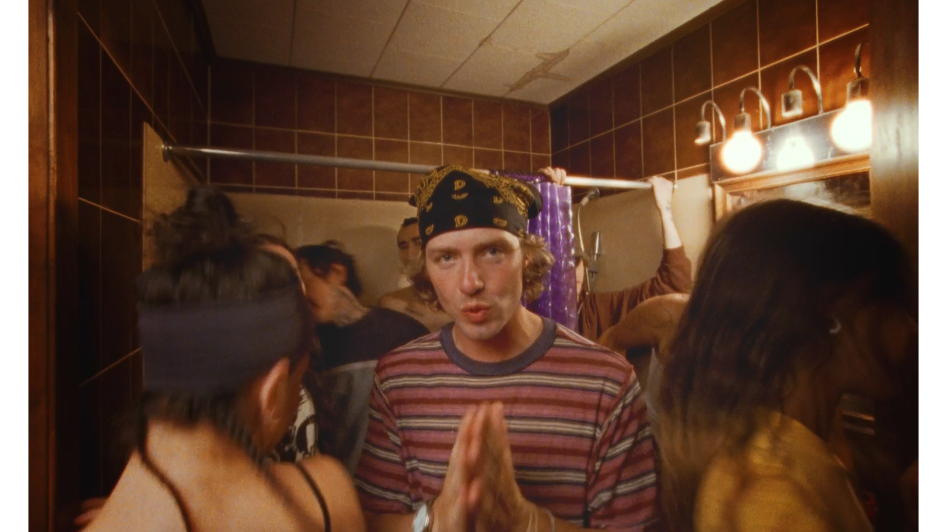 A man with a bandana on his head and a striped shirt stands in a crowded bathroom with several people around him. The bathroom has brown tiled walls, a purple shower curtain, and a mirror with four light bulbs above it. The man appears to be speaking
