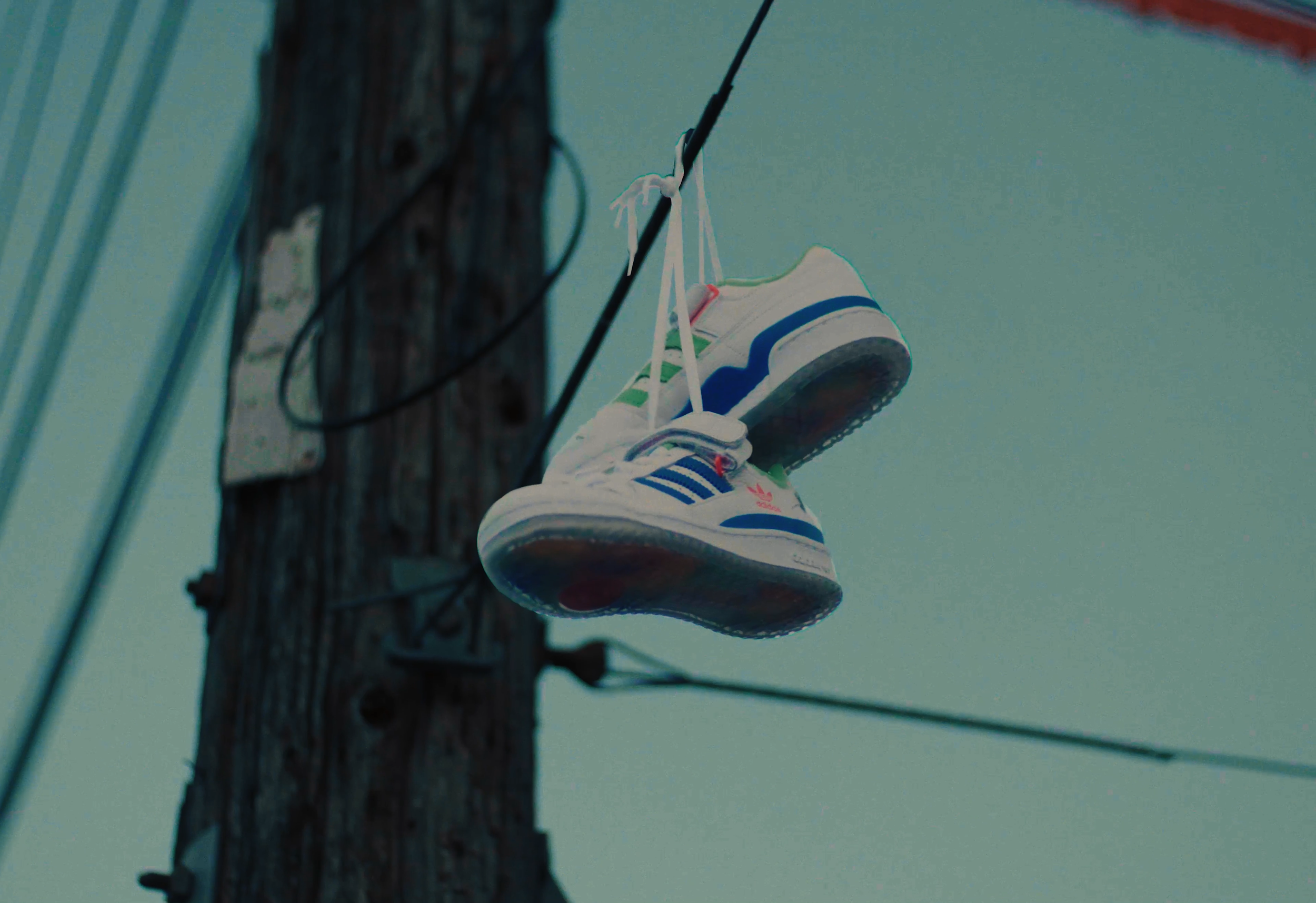 A pair of white Adidas sneakers with blue stripes and red accents hanging from a power line on a wooden utility pole.