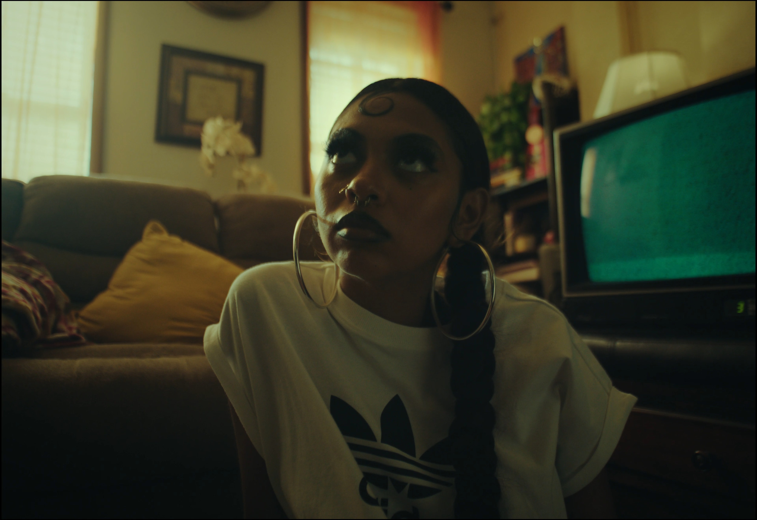 A woman with dark makeup and hoop earrings sitting on the floor of a living room with a TV, couch, and pillows behind her.