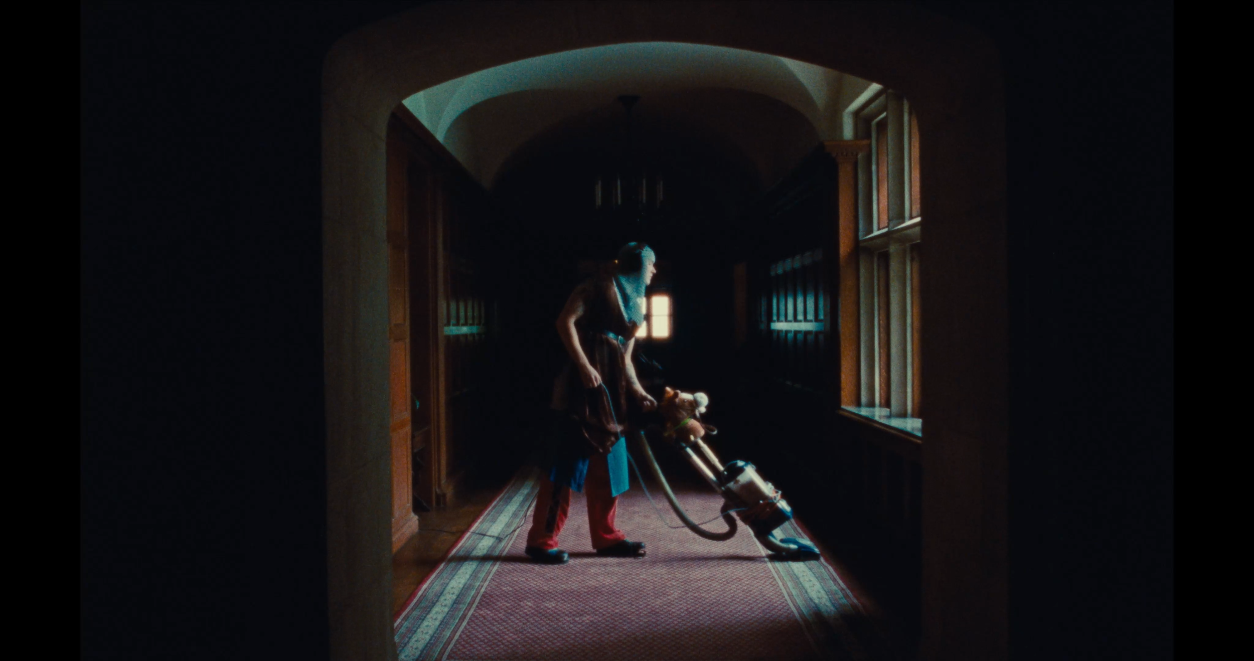 Person vacuuming an ornate hallway with wood-paneled walls and stained glass windows.
