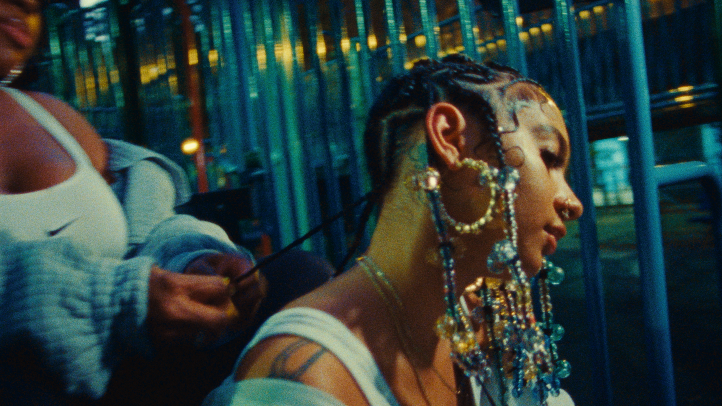 A woman with decorated, braided hair and multiple piercings gets her hair braided at a park or playground, with blue fences in the background.