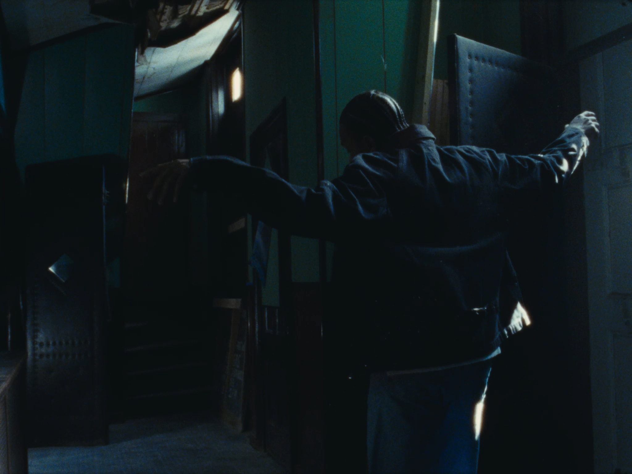 A person with cornrows extending from the crown to the nape is in a dark room with dim lighting. They are wearing a black jacket and jeans, with arms outstretched and touching walls or furniture, creating a silhouette against the low light.