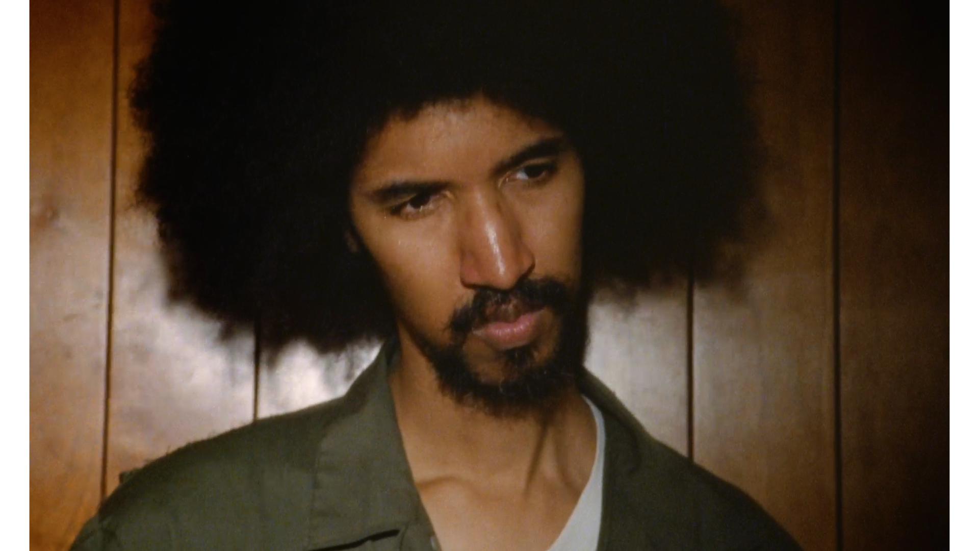 A man with a large afro hairstyle, a beard, and mustache, looking downward with a serious expression, standing in front of a wooden-paneled wall.