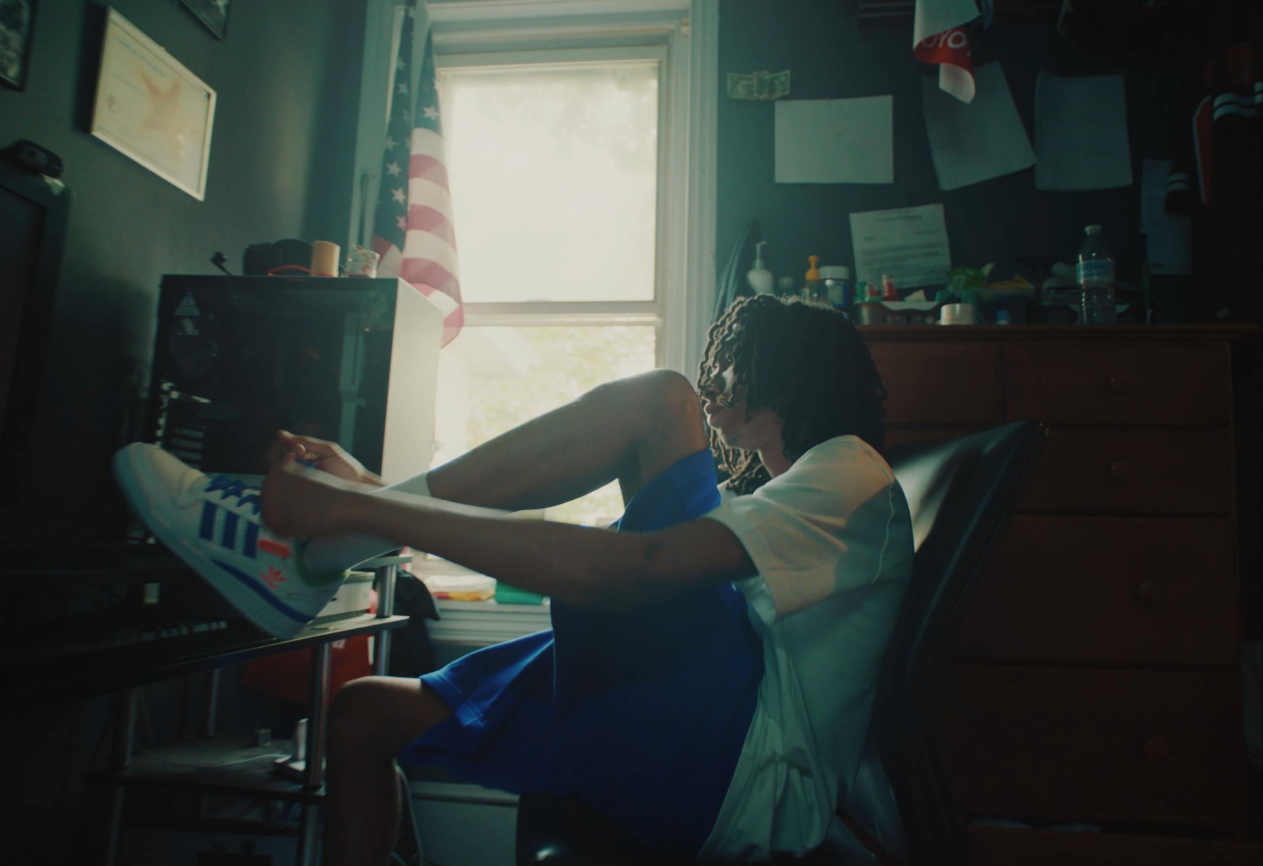A person with curly hair puts on a sneaker while sitting on a chair in a cluttered room with a window letting in sunlight.