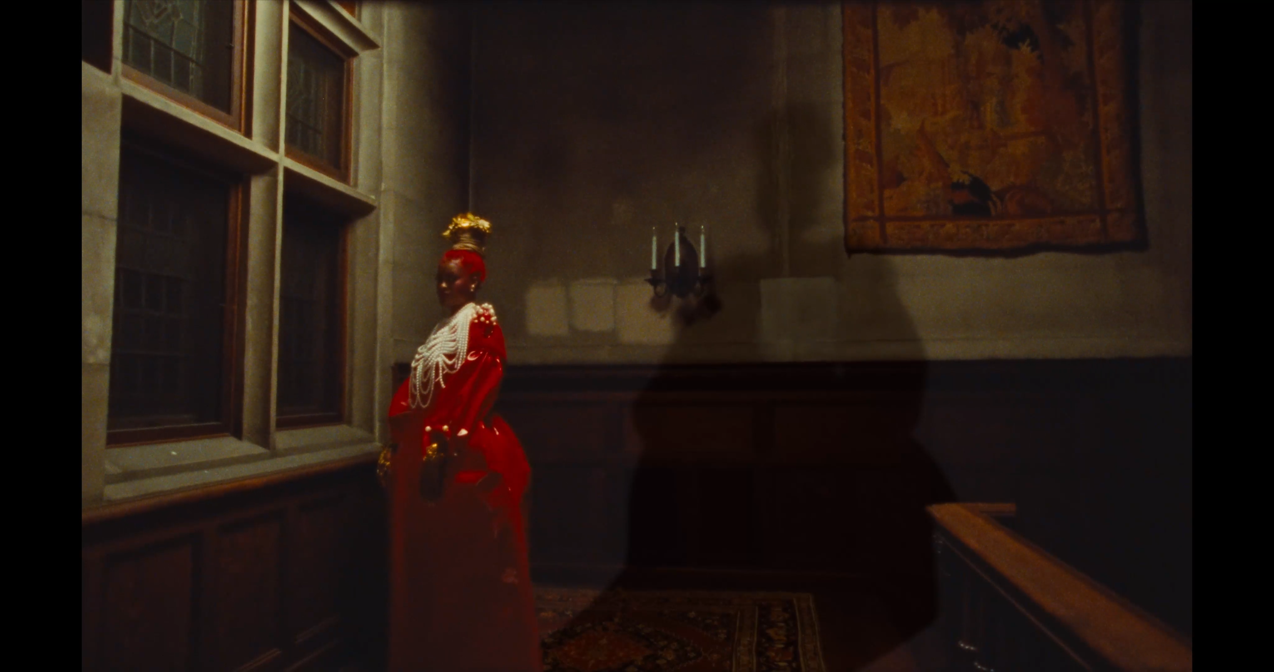 A woman dressed in a red gown with white and gold embellishments, wearing multiple layered pearl necklaces, stands in a dimly lit room near a window with wood framing, a tapestry on the wall, and a wall-mounted candle holder.