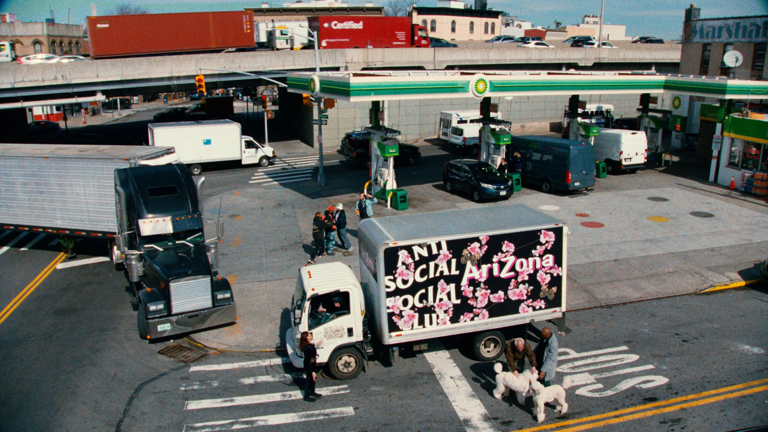 A busy city street scene with a BP gas station in the background, several parked cars, walking pedestrians, and a truck with decorated text and flowers on its side.
