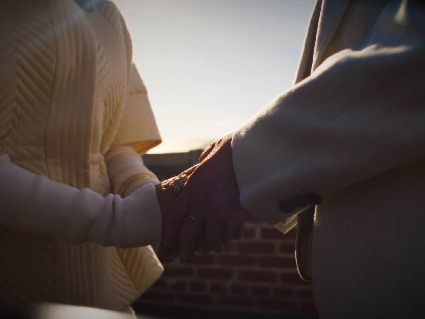 Two people holding hands, with sunlight shining in the background, standing outdoors near a brick wall.