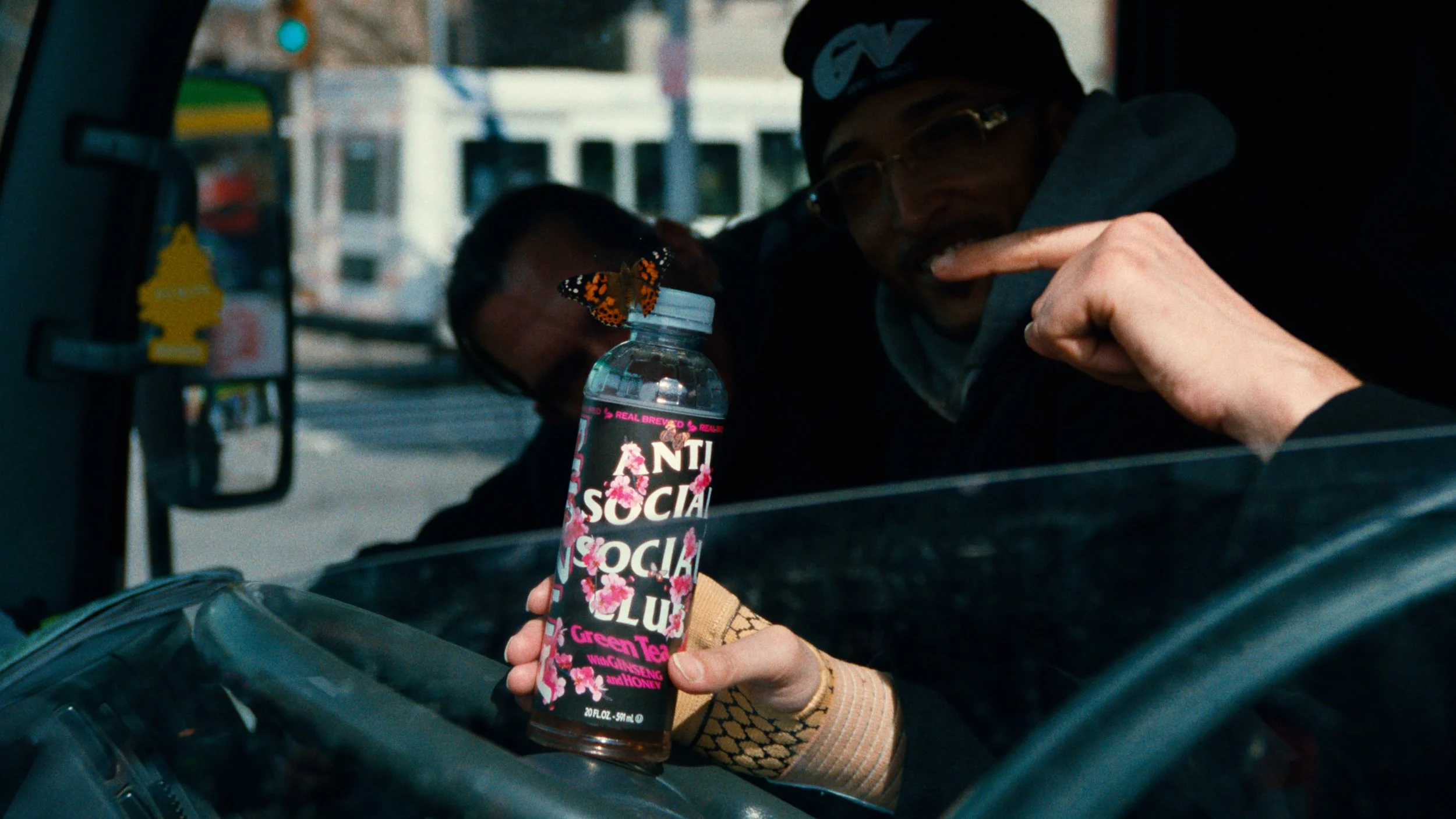 A person holding a bottle labeled 'Anti Social Social Club Green Tea' with a butterfly perched on the cap, inside a vehicle with two other people visible in the background.