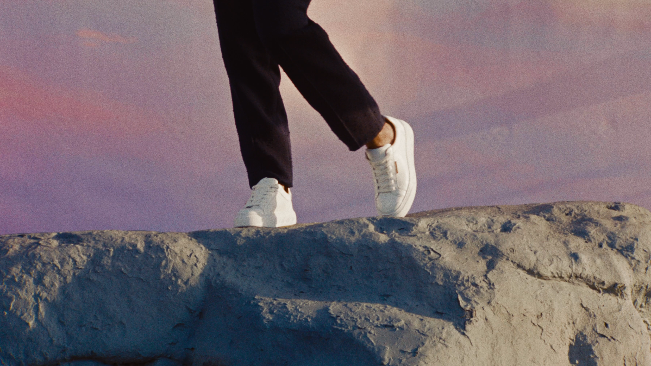 Close-up of a person wearing black pants and white sneakers walking on a rocky surface with a colorful sky in the background.