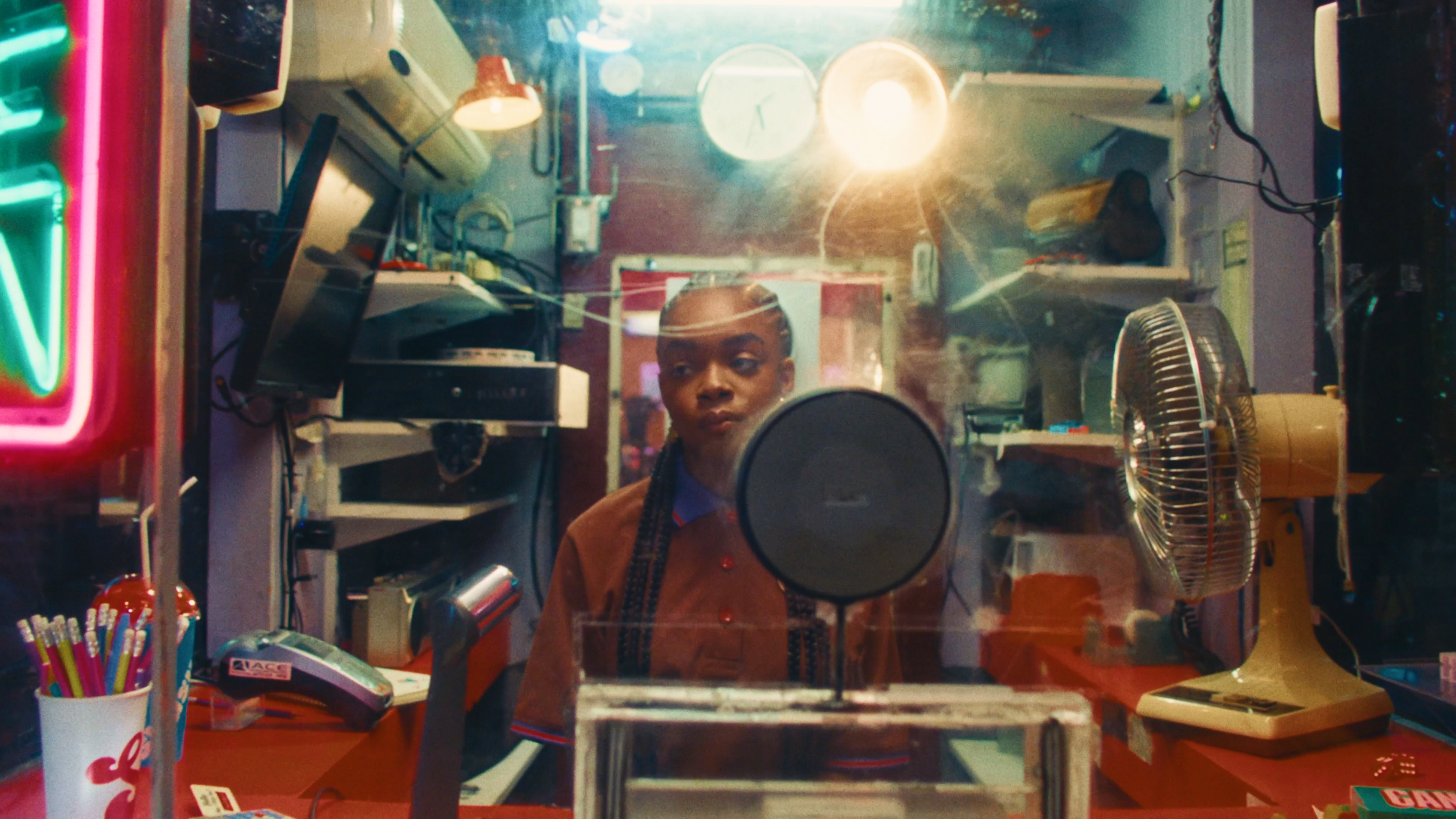 Person standing behind a counter in a cluttered room viewed through a glass window with a large round object in the foreground.