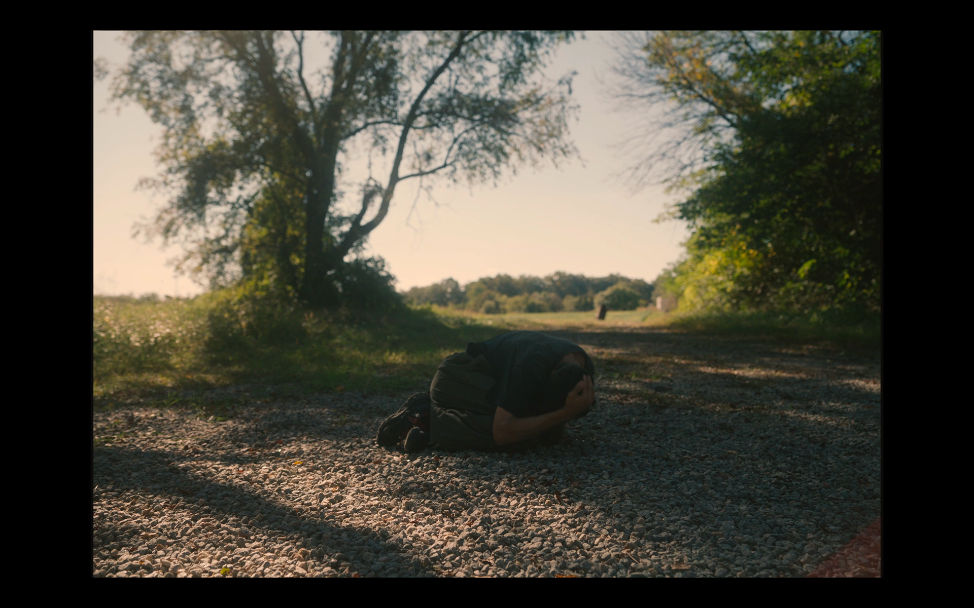 A person is kneeling on a gravel path outdoors, holding their head in distress or despair, during daylight with trees and greenery around.