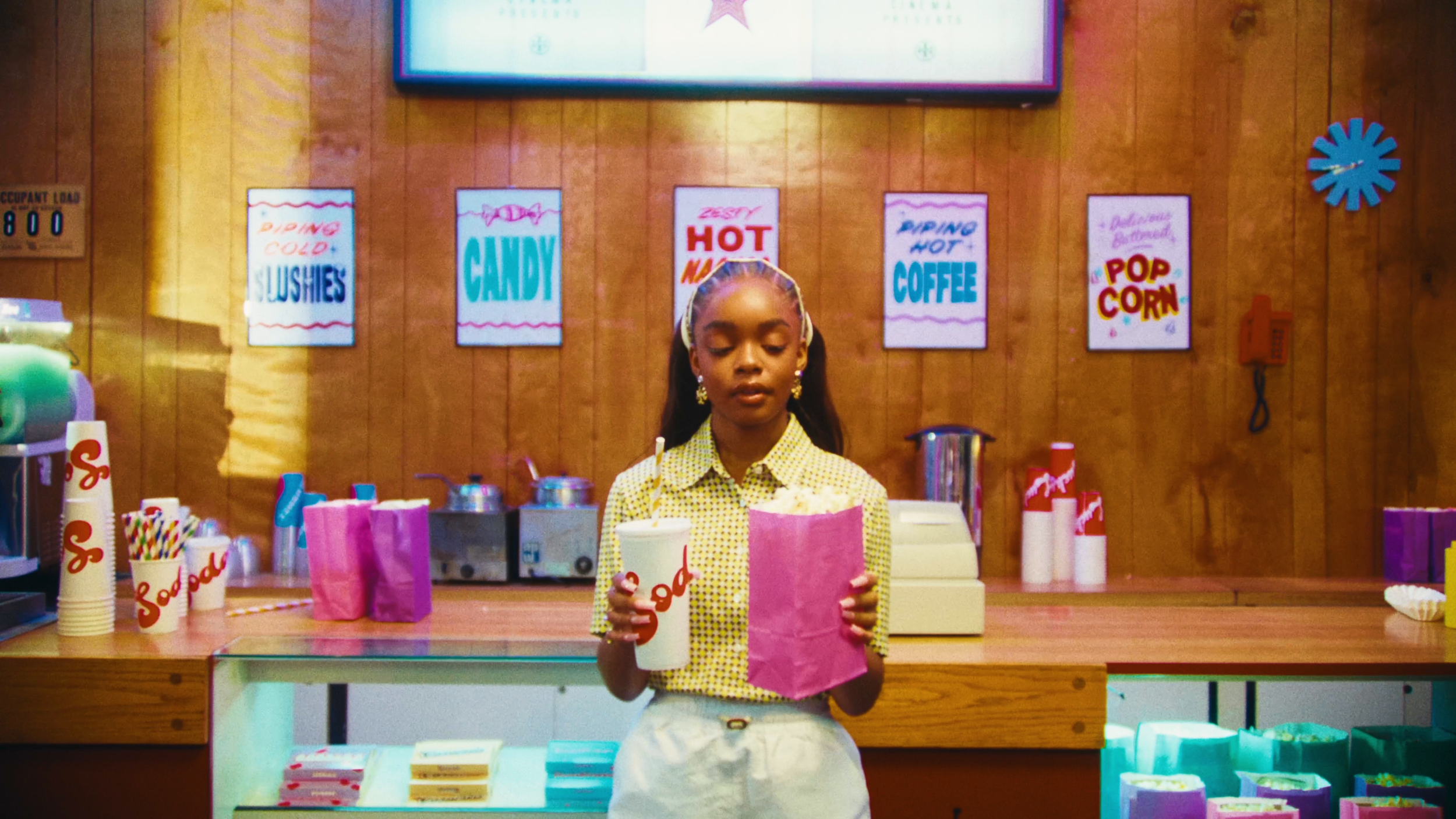 A girl standing behind a counter in a snack shop, holding a paper cup with a straw and a pink paper bag. The shop has colorful signs advertising various treats like candy, popcorn, and coffee on a wooden wall.