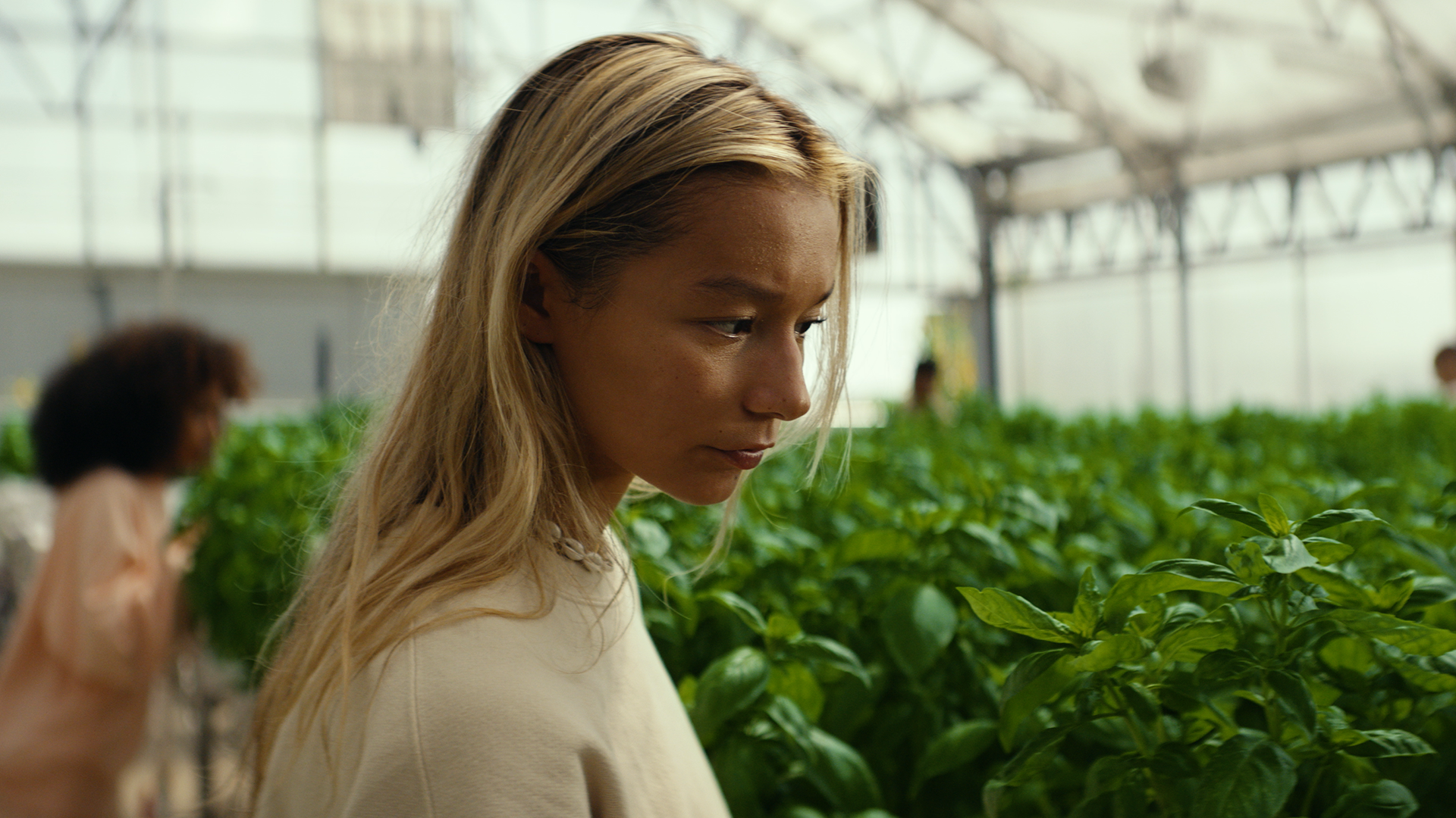 A young woman with blonde hair looking at green plants in a greenhouse.