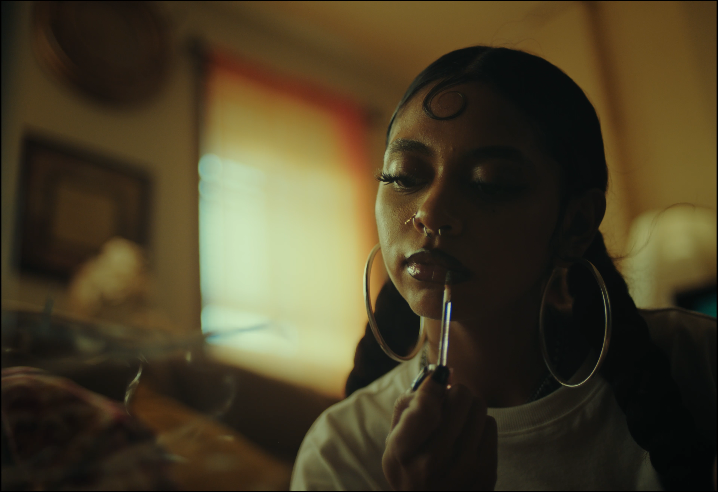 A young woman with dark hair in a braid, large hoop earrings, and a septum piercing, applies lipstick while looking into a mirror in a dimly lit room.