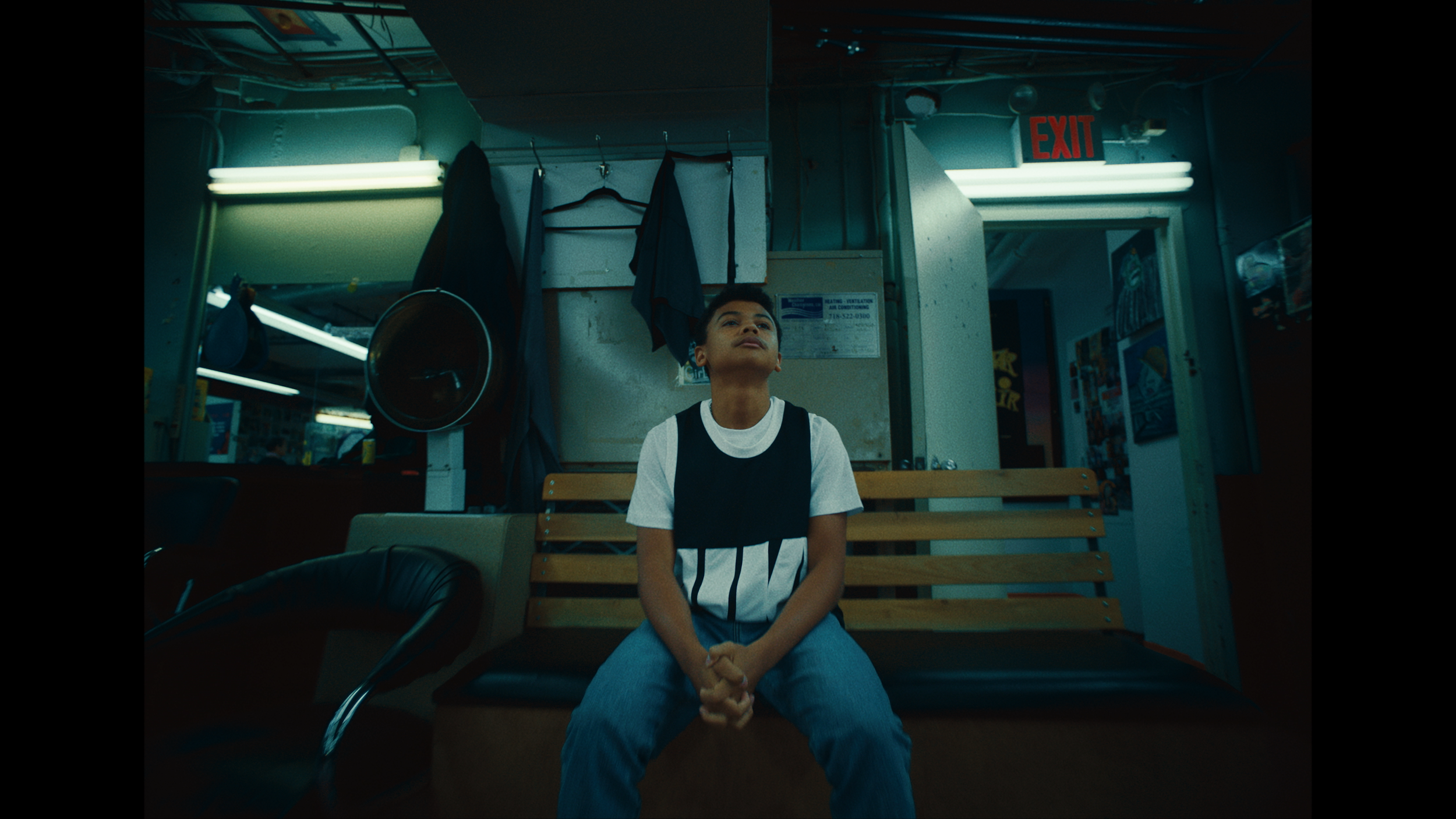A young boy sitting on a wooden bench inside a laundromat, looking upward with hands clasped, surrounded by washing machines, hanging clothes, and fluorescent lighting.