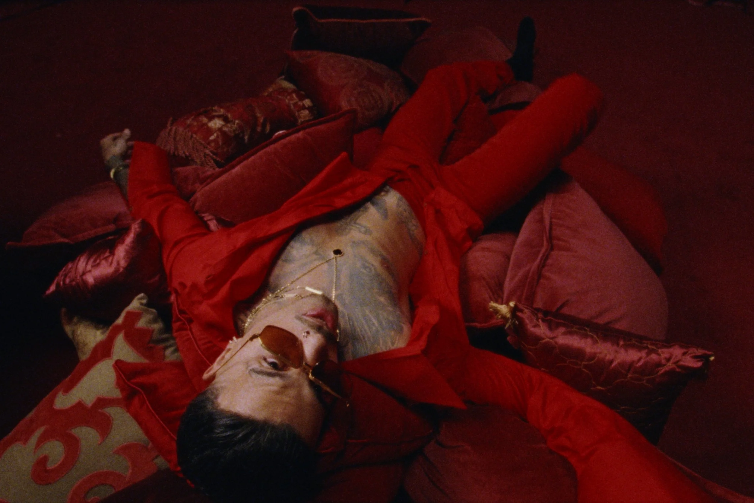 A person lying on a bed surrounded by red and burgundy pillows, wearing sunglasses and jewelry, with a relaxed pose and a bluish tattooed chest.