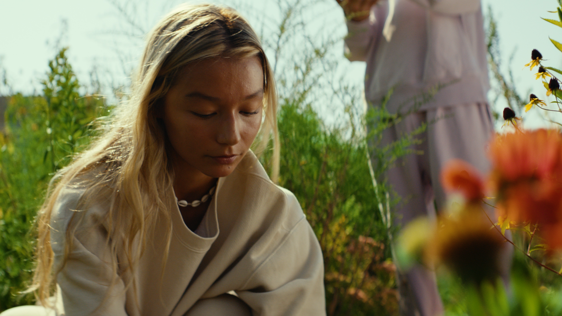 A young woman with long blonde hair, wearing a beige hoodie and shell necklace, looks down thoughtfully while surrounded by greenery and flowers outdoors.