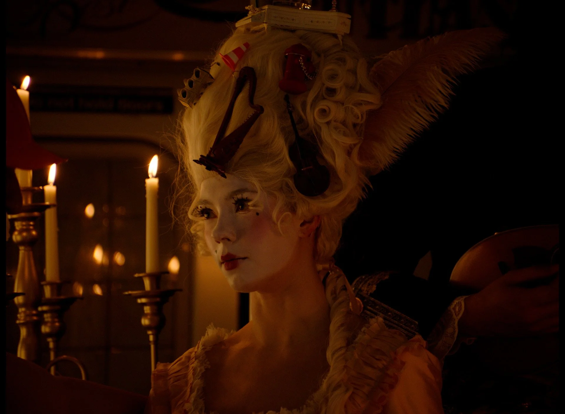A woman dressed in elaborate historical costume with a tall, curly, blond wig adorned with hairpieces and accessories, illuminated by candlelight with candles in the background.