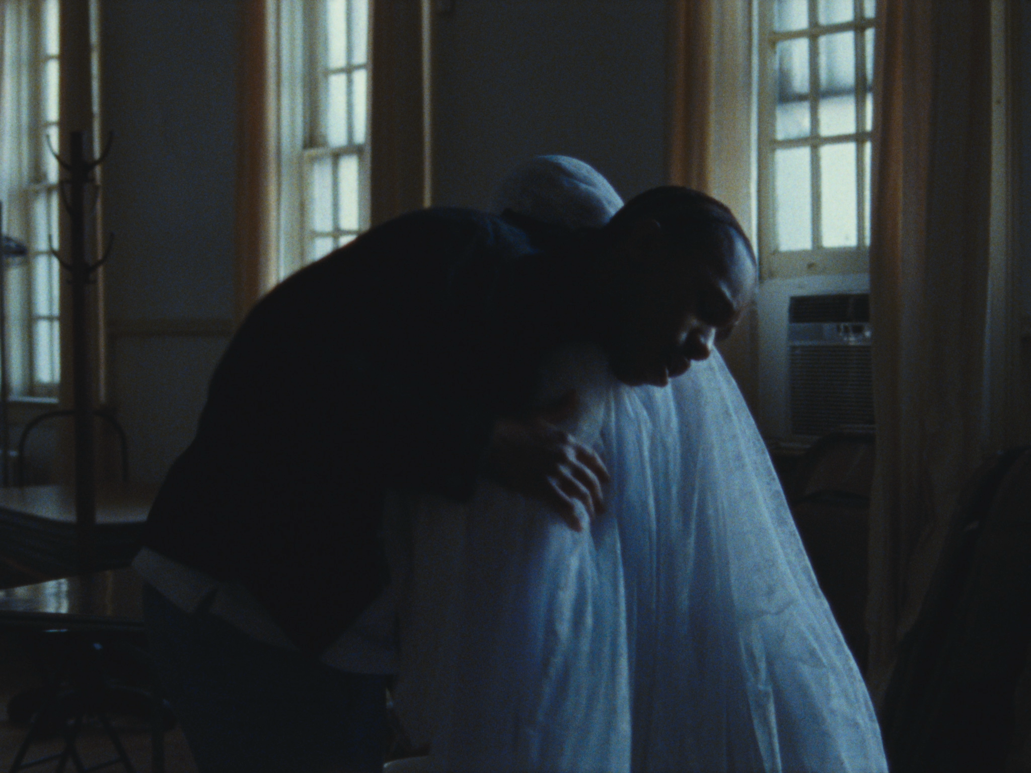 A person leaning over and hugging someone wrapped in a white sheet or gown, in a dimly lit room with large windows and curtains.