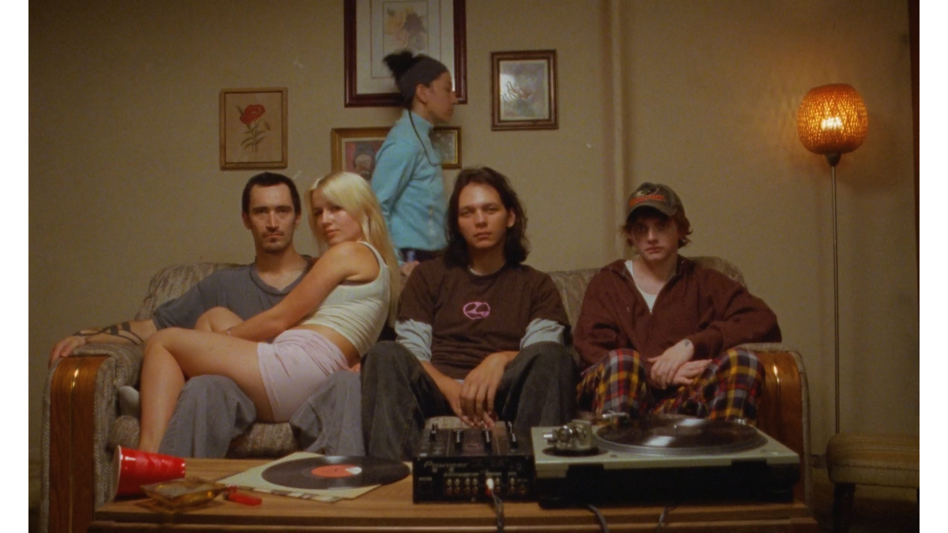 Four teenagers sitting on a couch with a turntable and records on a wooden table in front of them, and a woman walking by in the background. The room has framed pictures on the wall and a tall floor lamp with a woven shade.