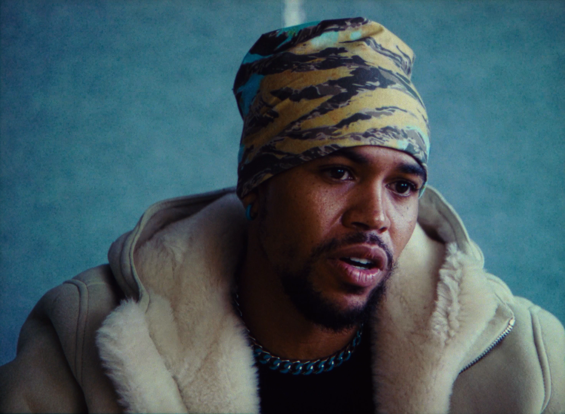 A man wearing a camo head wrap, tan jacket with fur collar, and a chain necklace, appears to be speaking or reacting with a serious expression.