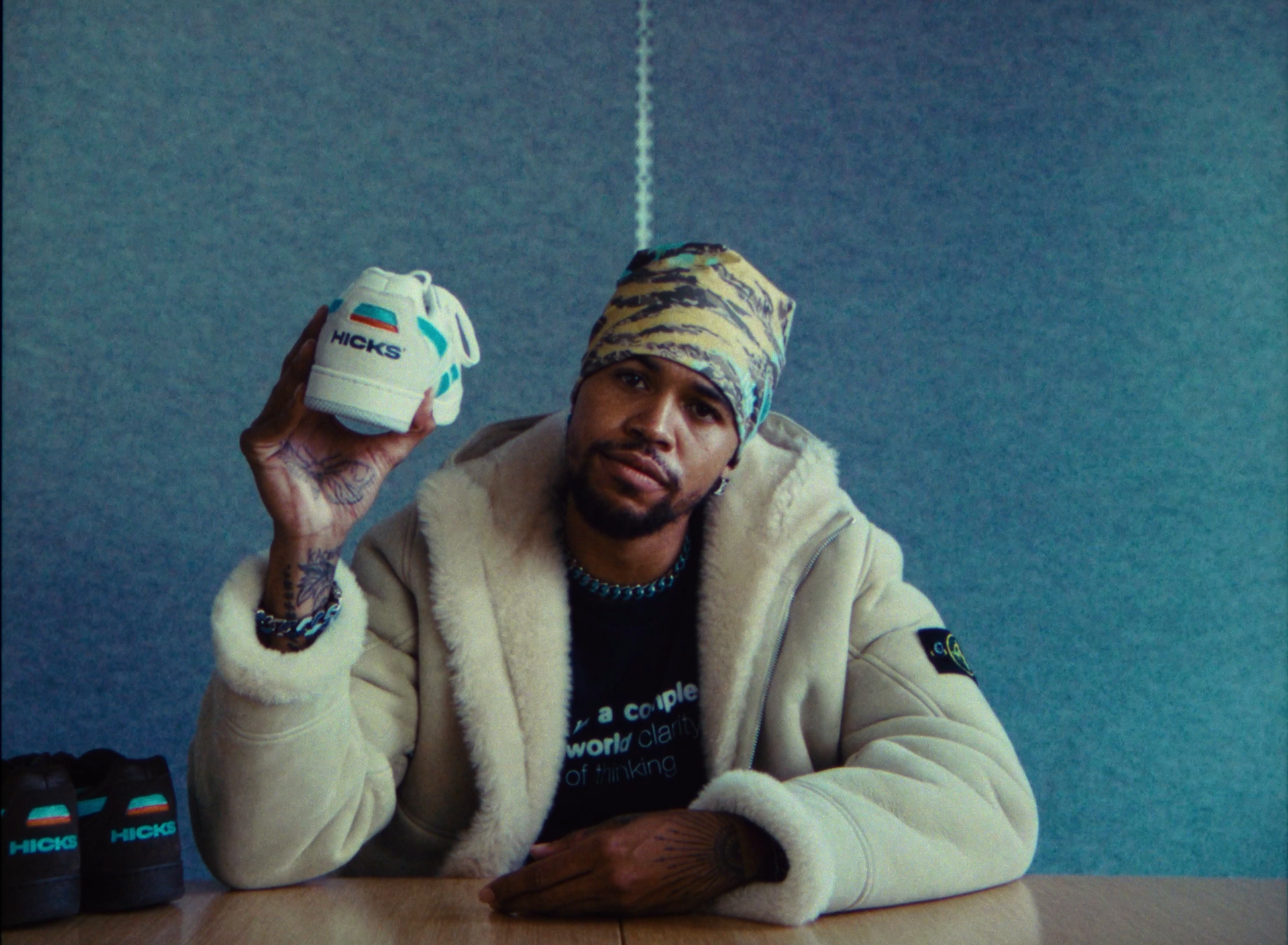 A man with tattoos and a patterned bandana on his head sitting at a table, holding a white sneaker with the word 'HICKS' and a flag on it, with more sneakers on the table.